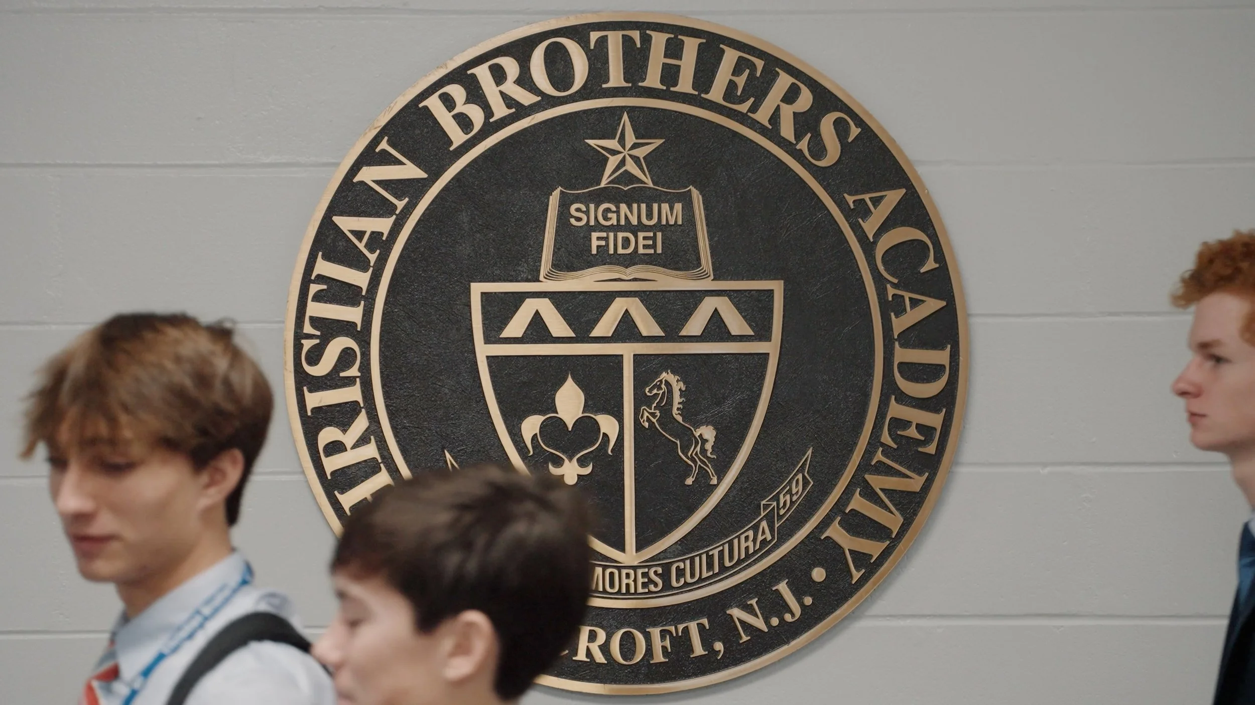Three students walking in front of the Princeton University seal on a gray wall. The seal features a shield with a star, an open book with the Latin phrase 'Signum Fidei,' and symbols of a fleur-de-lis and a horse.