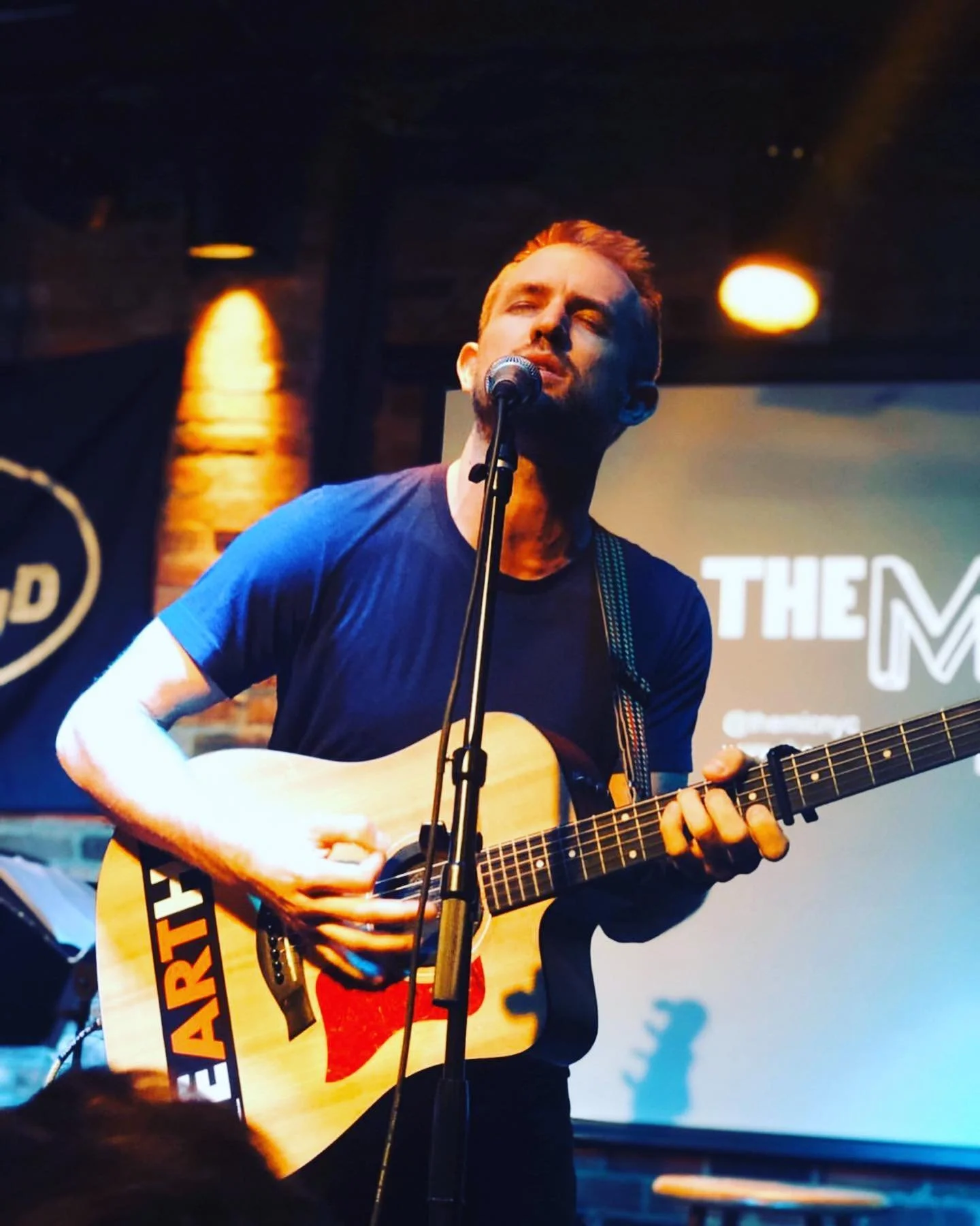 A man performing on stage with eyes closed, playing an acoustic guitar and singing into a microphone, illuminated by stage lights, in a dimly lit venue.