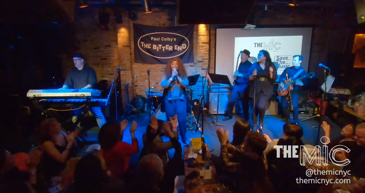 A band performing on stage in a brick-walled venue with a sign that reads 'Paul Colby's The Bitter End'. A woman is singing at the center, and a guitarist, keyboardist, and backup singer are also performing. Audience members are clapping and enjoying