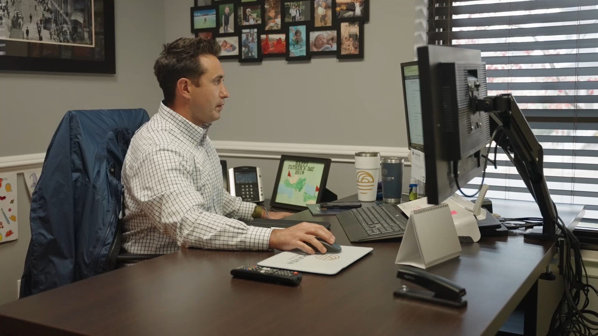 A man sitting at a desk working on a computer in an office, with framed photographs on the wall and a window with blinds behind him.