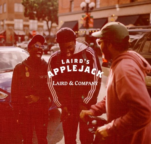 Three people standing outside on a street during daytime, with buildings and parked cars in the background. They appear to be engaged in conversation, with one person wearing a cap and glasses, and the others in casual attire.