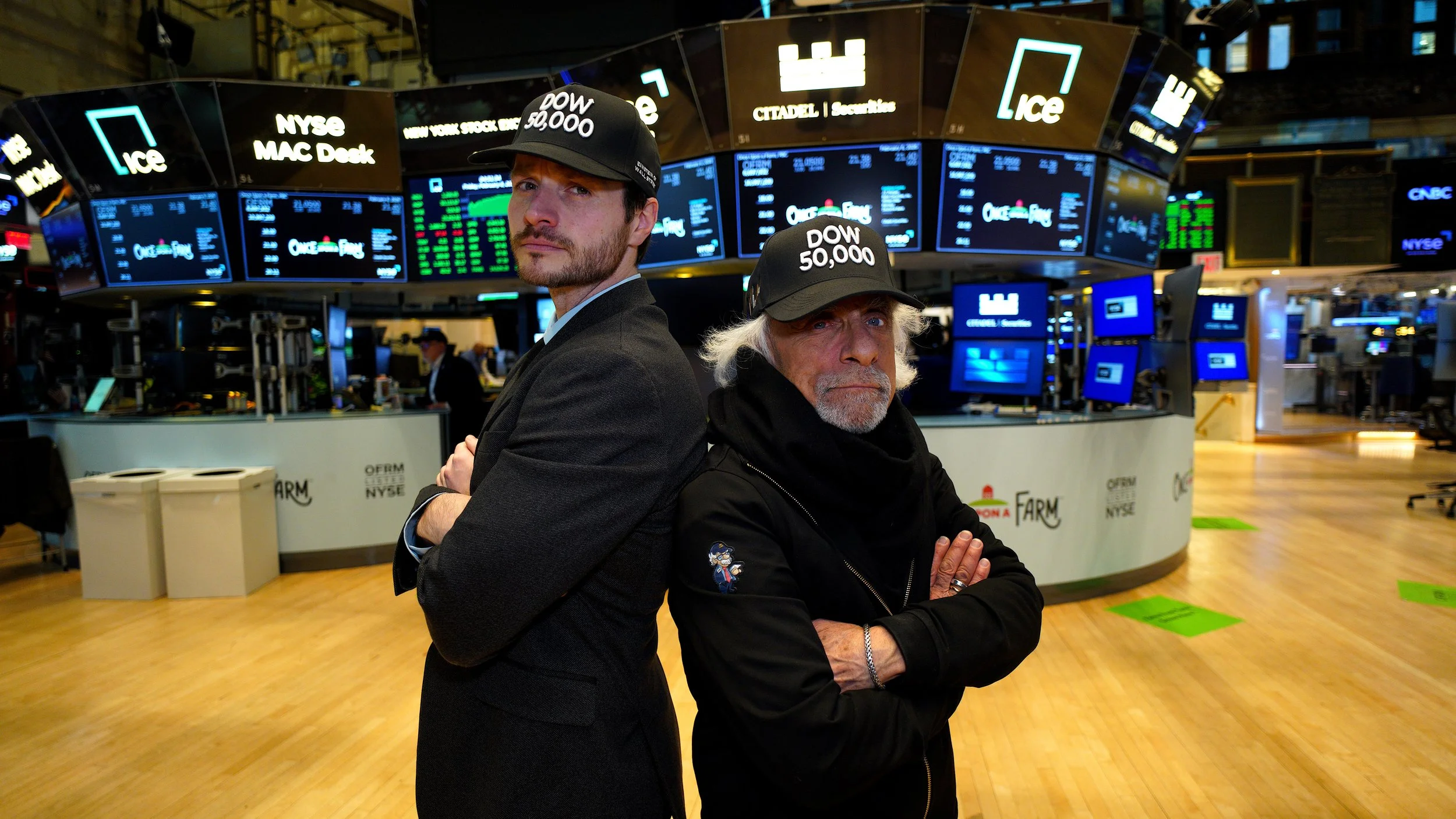 Two men stand back-to-back on the trading floor of a stock exchange with their arms crossed, wearing black caps with political and financial slogans, amidst electronic stock market displays.