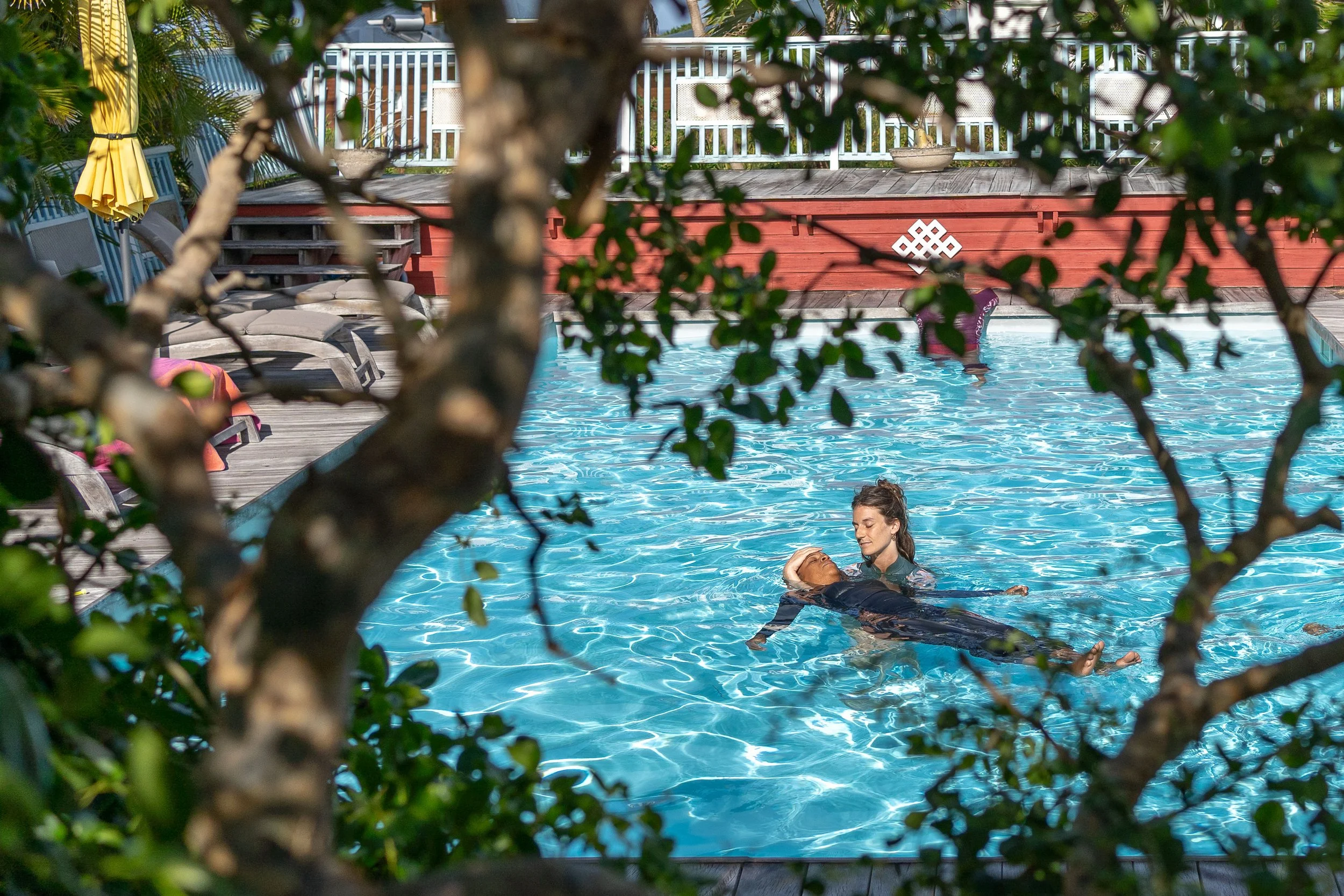 Piscine avec deux personnes nageant, vue partielle à travers des branches d'arbre avec des feuilles vertes, clôture blanche en arrière-plan, parapluie jaune plié, fauteuils de piscine, deck en bois.
