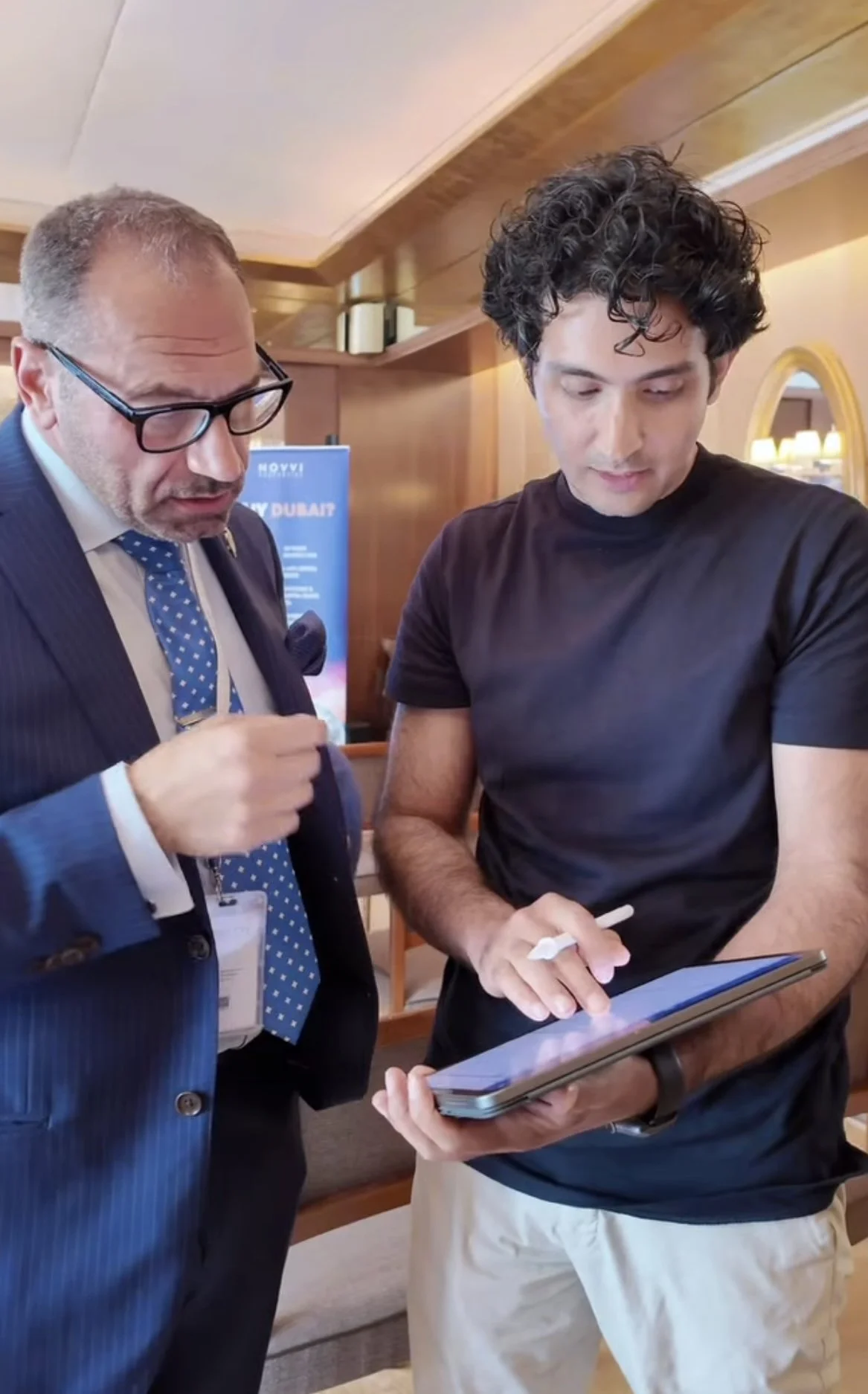 Two men, one dressed in a business suit and the other in casual clothing, are engaged in a conversation and looking at a tablet device together in a warmly lit indoor setting.