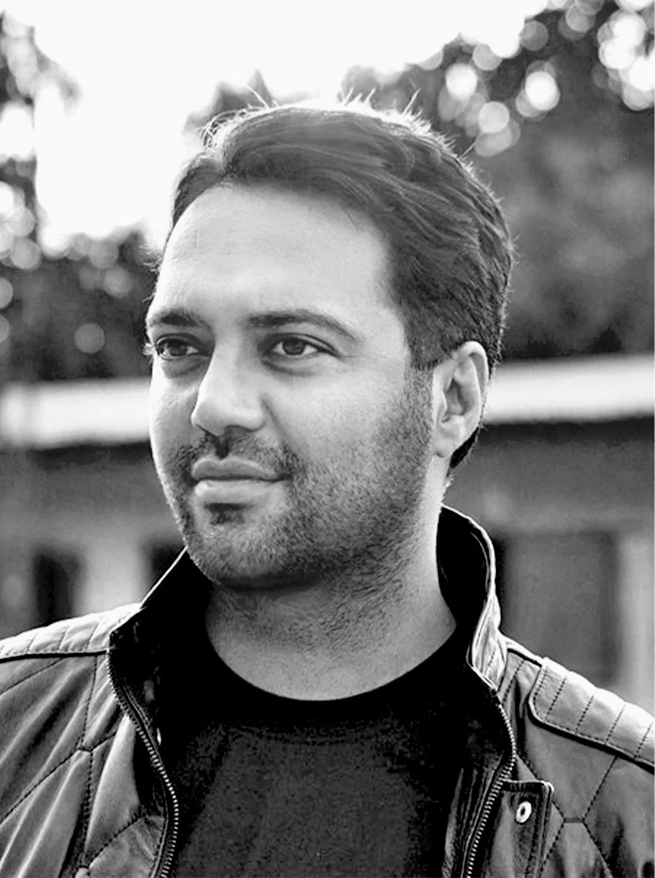 Black and white portrait of a young man with styled hair, light facial hair, and wearing a jacket with a quilted pattern, outdoors with blurred trees in the background.
