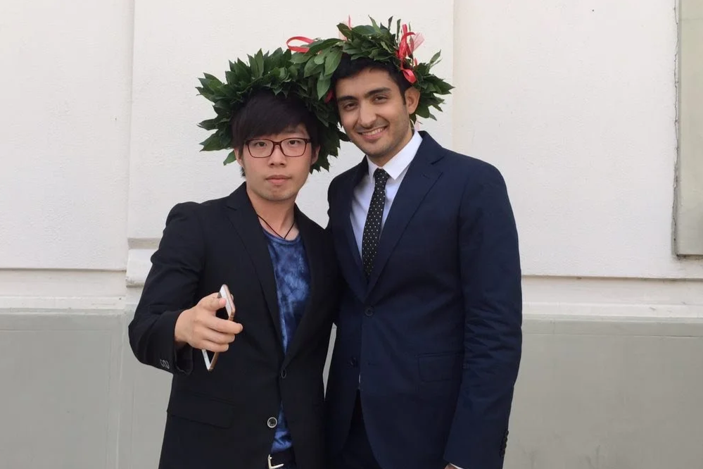 Two men in suits standing side by side wearing wreaths made of green leaves on their heads, smiling at the camera.