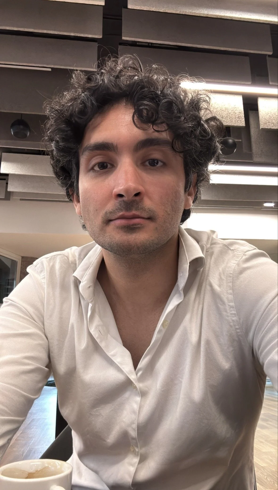 A man with curly black hair wearing a white button-up shirt, sitting at a table in a modern indoor setting with acoustic panels on the ceiling and a coffee cup in the foreground.