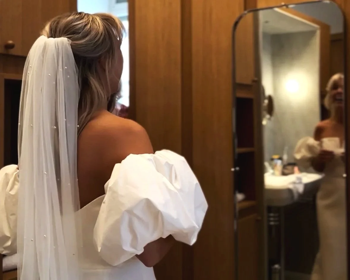 A bride with a wedding dress and veil is looking at her reflection in a mirror, smiling, inside a wooden-paneled room.