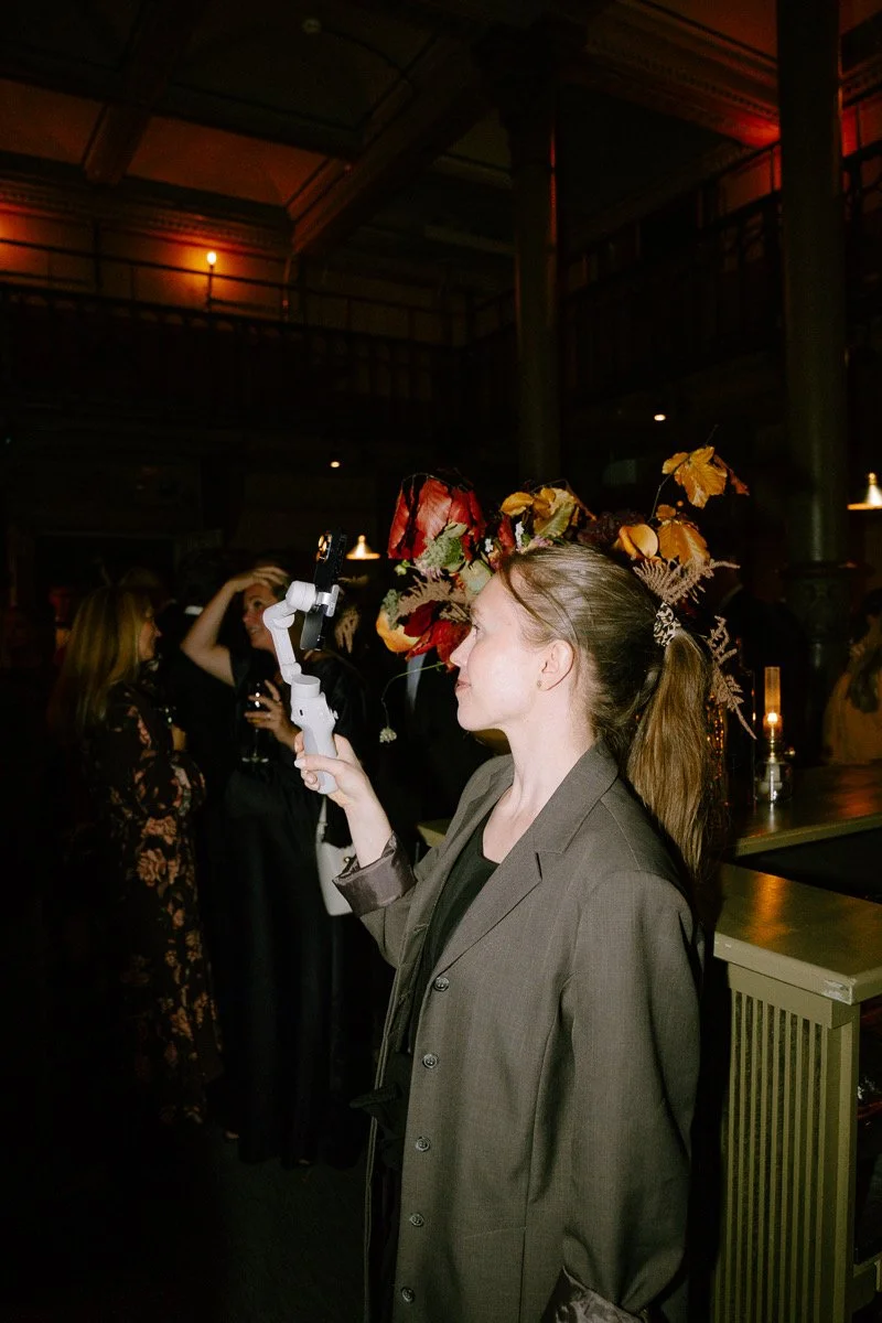 A woman wearing a gray blazer stands in a dimly lit venue, holding a small camera or device. She has a large floral headpiece with autumn leaves and red flowers. In the background, other women are seen talking and holding drinks, with warm lighting and industrial-style architecture.
