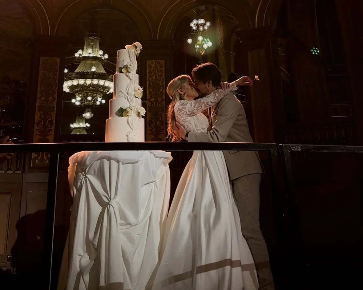 A bride and groom sharing a kiss at their wedding reception, with a tall wedding cake decorated with white flowers in the background.