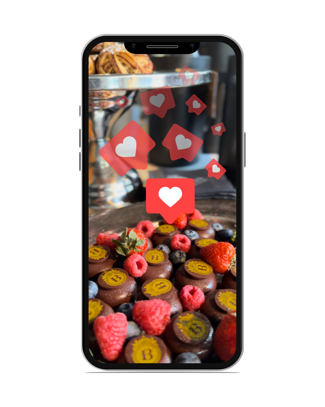A smartphone displaying a photo of assorted chocolates topped with strawberries, raspberries, and blueberries, with floating social media 'like' icons overlaid.