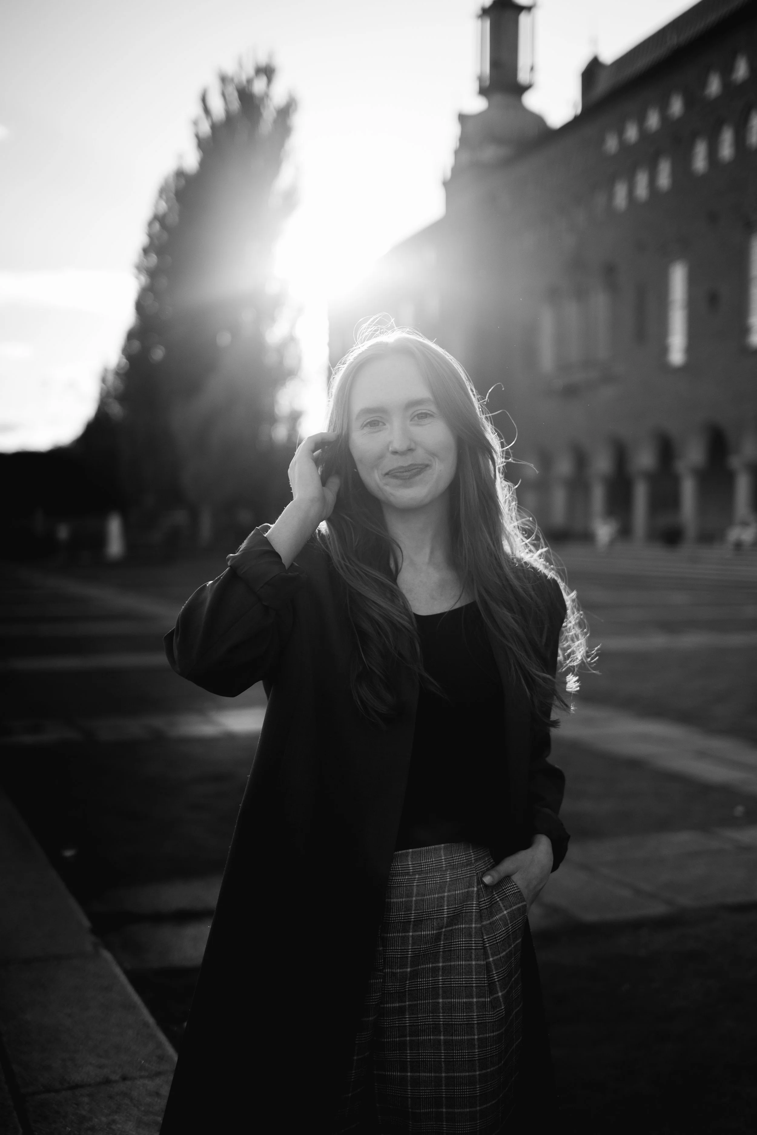 A woman with long hair smiling at the camera, standing outdoors with the sun setting behind her, in front of historic brick buildings and trees.