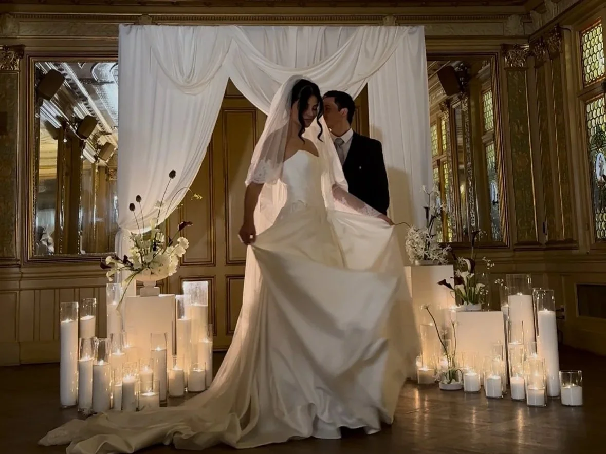Bride and groom posing on a wedding stage with white draped curtains, flowers, and candles around.