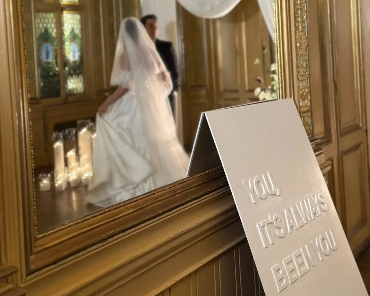 A bride and groom are reflected in a mirror, with the bride wearing a white wedding dress and veil, standing next to the groom in a formal outfit. A white sign with embossed text reading 'YOU IT'S ALWAYS BEEN YOU' leans against the mirror in the foreground, in a room with wood-paneled walls and candles.
