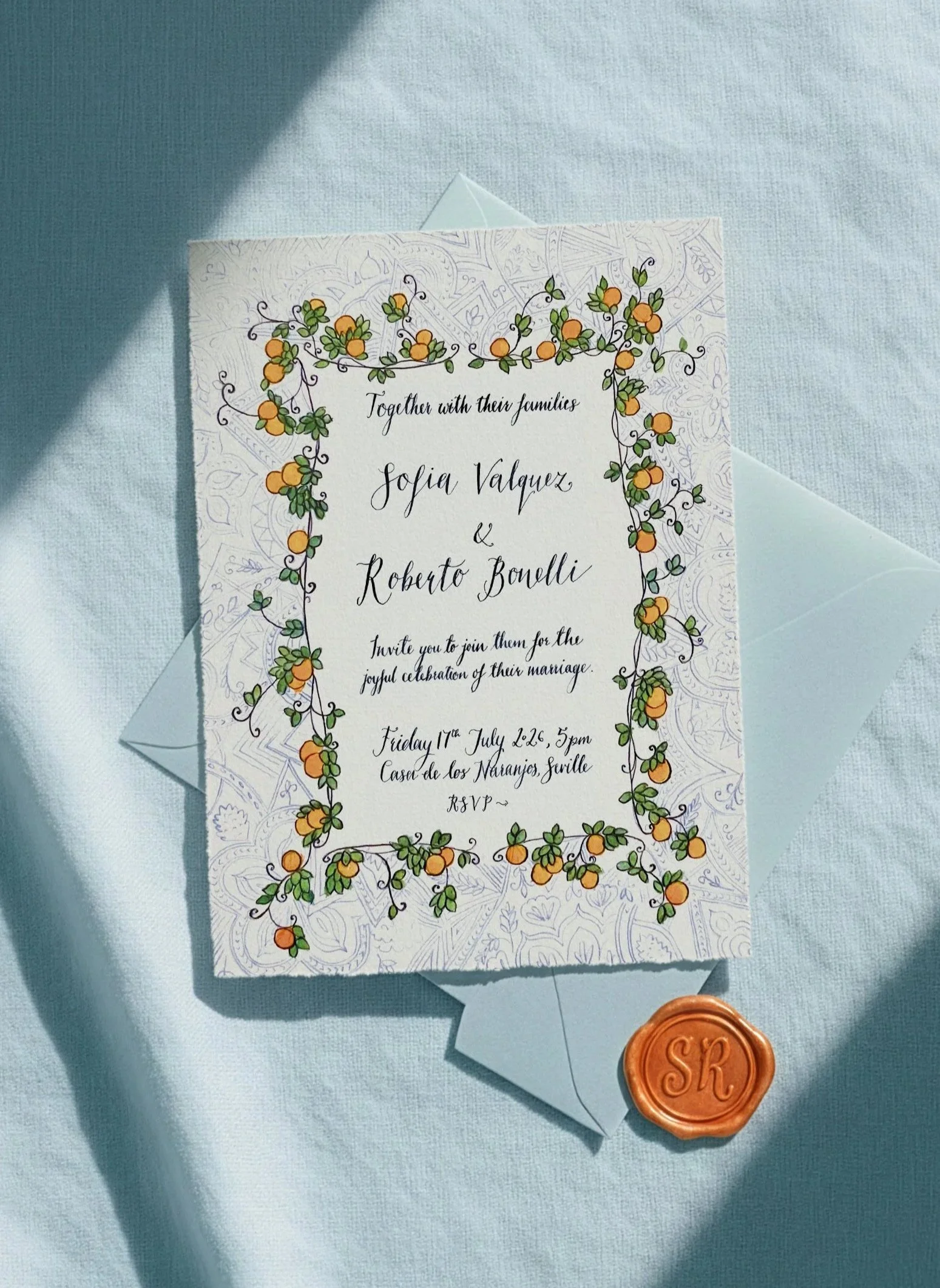 Wedding invitation card with a floral border featuring orange and green flowers, placed on a light blue fabric surface with a matching light blue envelope and a wax seal embossed with the initials 'SR'.