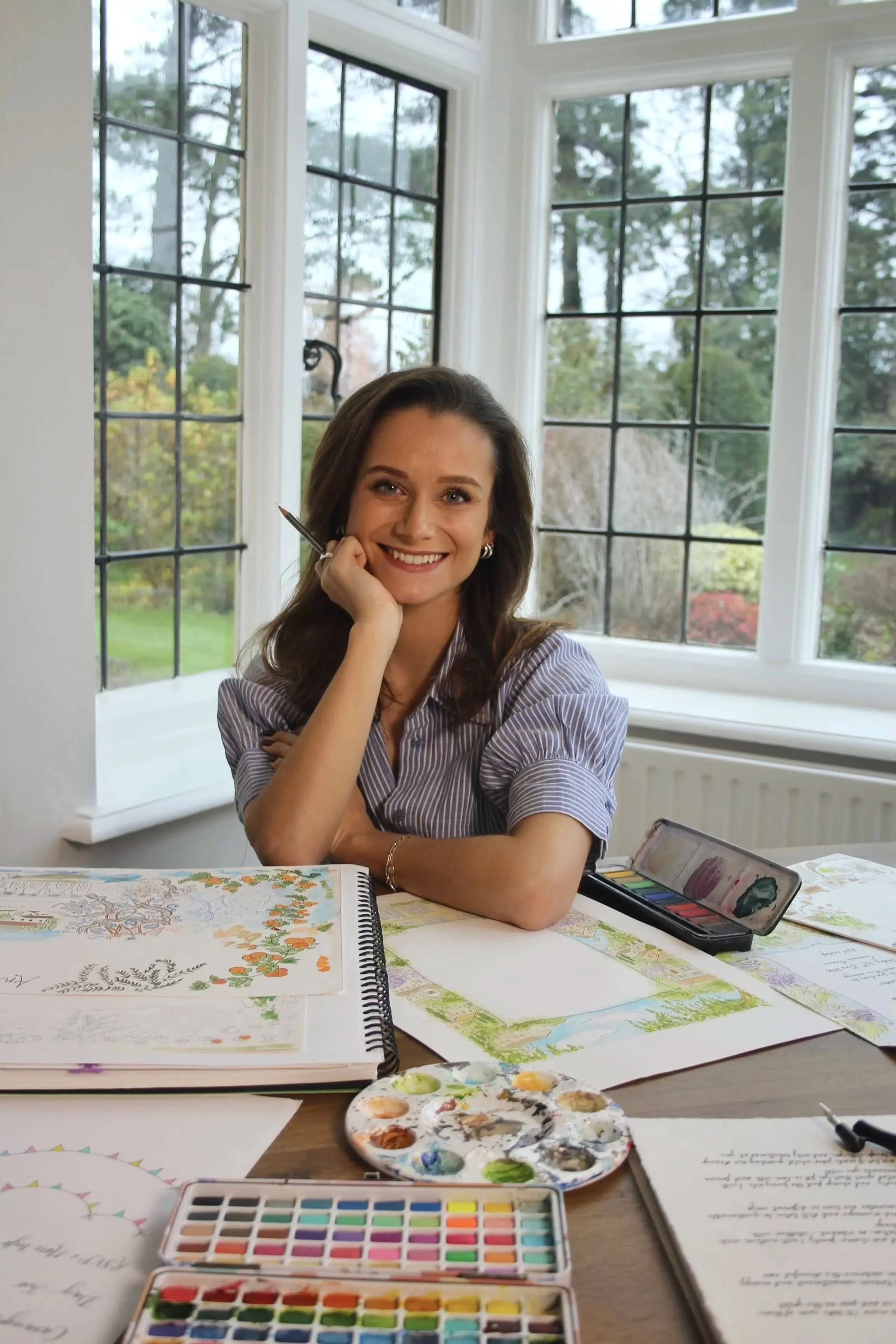 A woman with shoulder-length brown hair, smiling, sitting at a table with watercolor paintings and art supplies, near large windows with a view of trees outside.