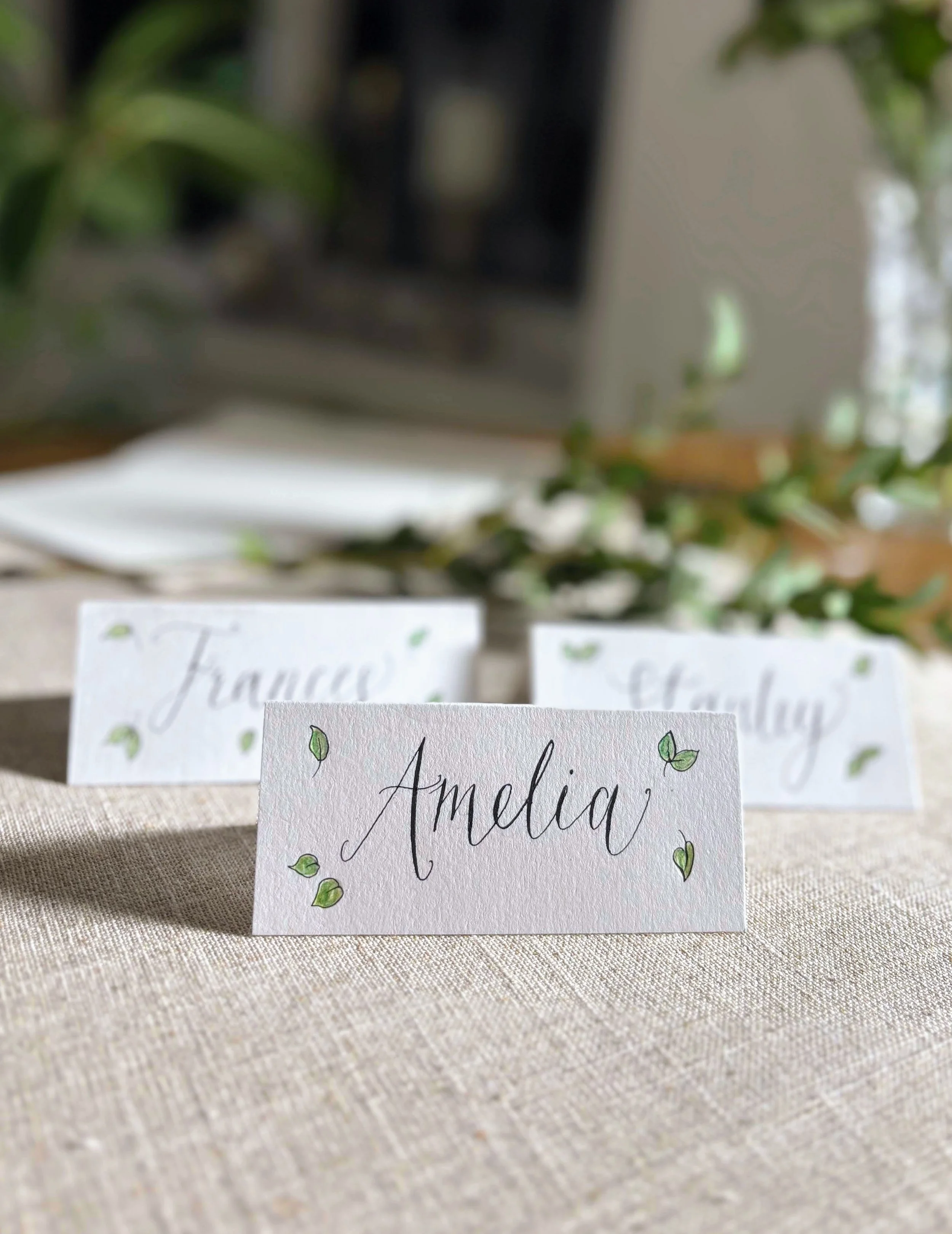 Name card with the word 'Amelia' written in elegant cursive, decorated with small green leaves, placed on a beige tablecloth at an event or wedding.