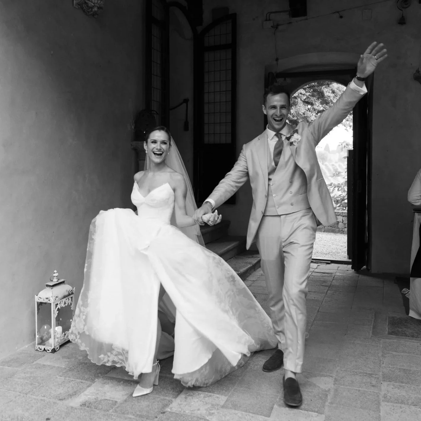 A bride and groom are walking together, smiling and holding hands, inside a building with open door and window, during their wedding celebration.
