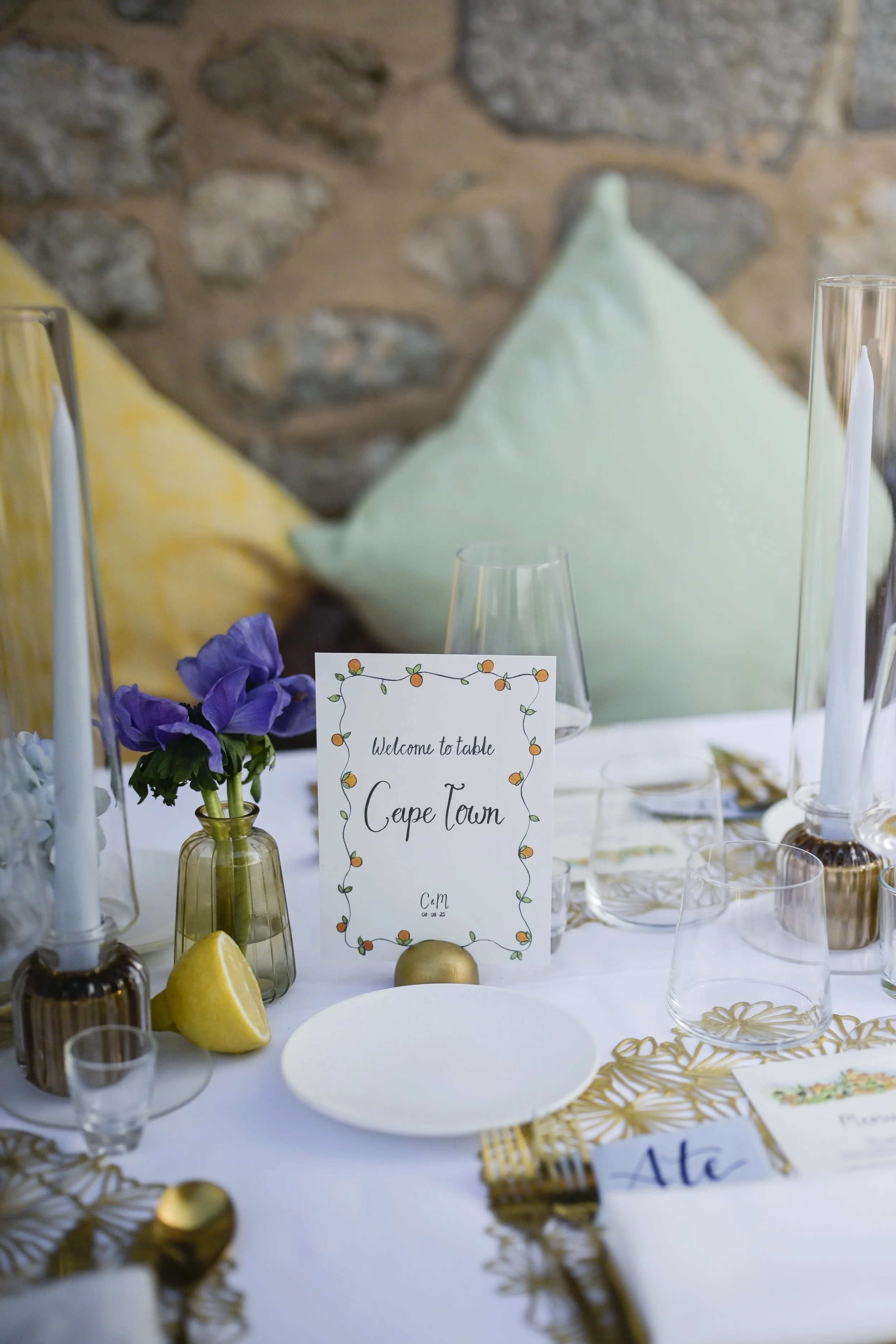 A decorated dinner table with a welcome sign, purple flowers in a small plantholder, candles, glassware, and gold-colored cutlery, against a background of a stone wall and pillows.