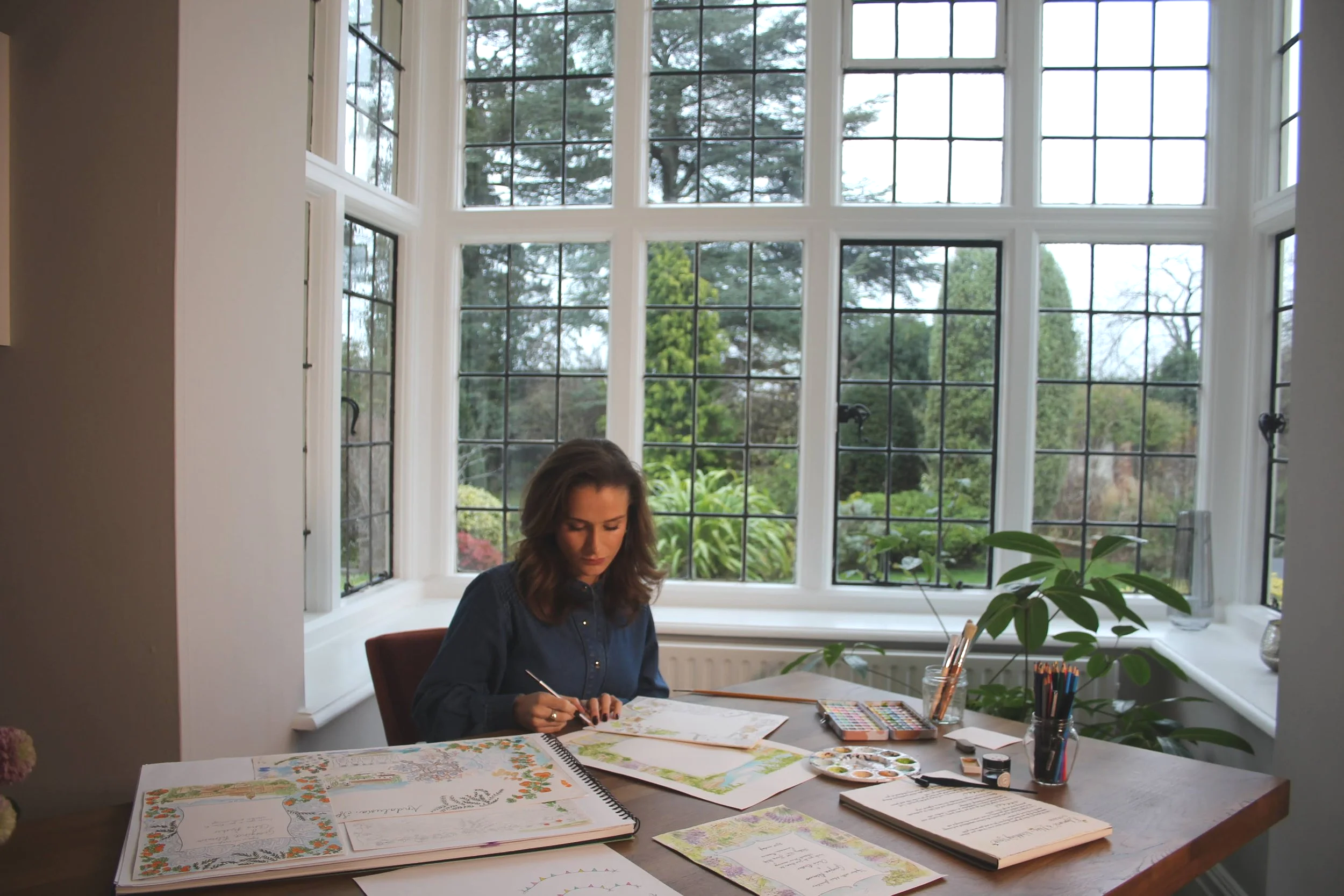 An artist is sitting at a wooden table in a bright room with large windows overlooking a garden. She is drawing or painting in a sketchbook with art supplies scattered on the table.