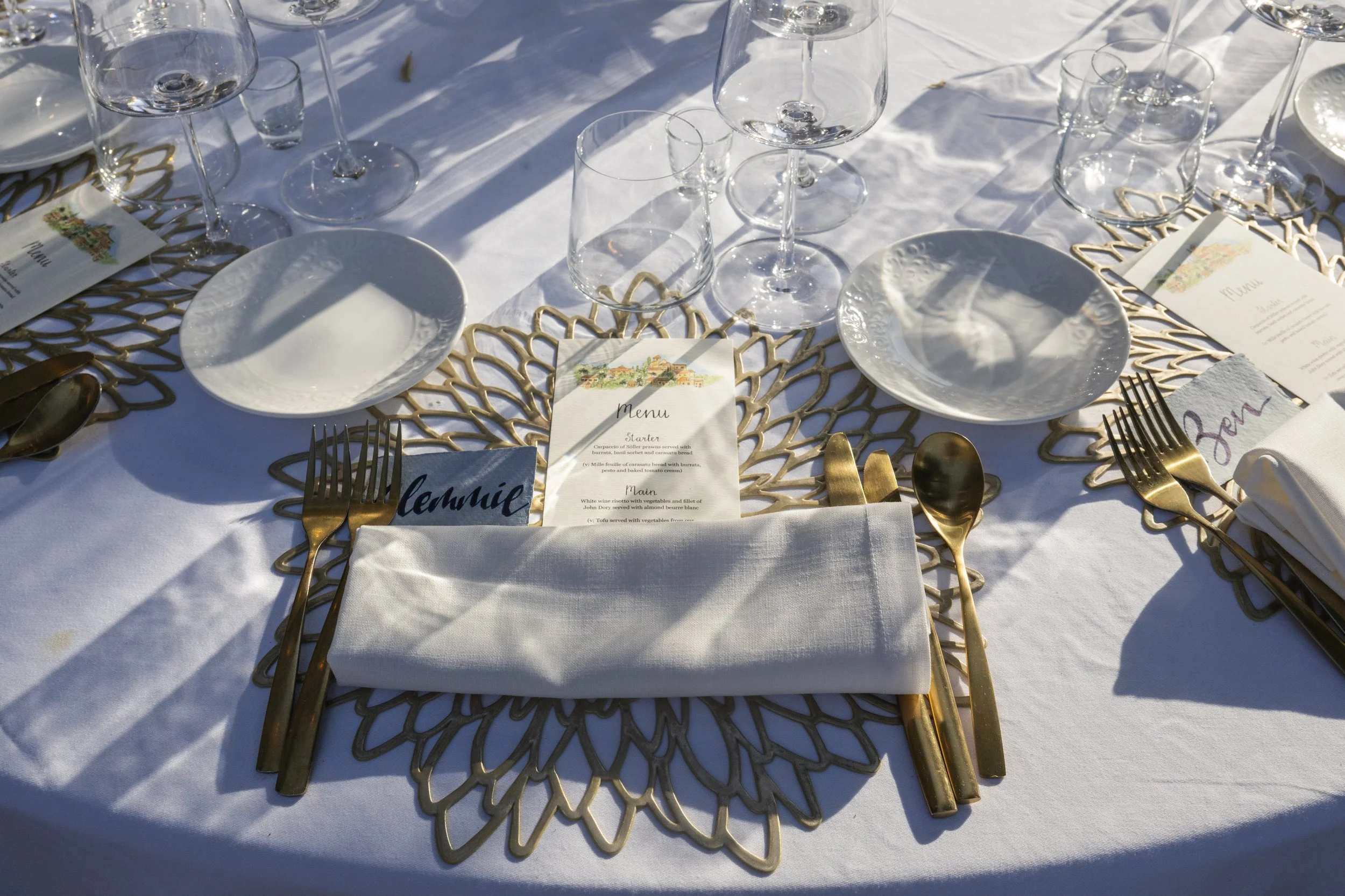 Elegant outdoor dining table set with gold cutlery, white napkin, glassware, and decorative placemats.