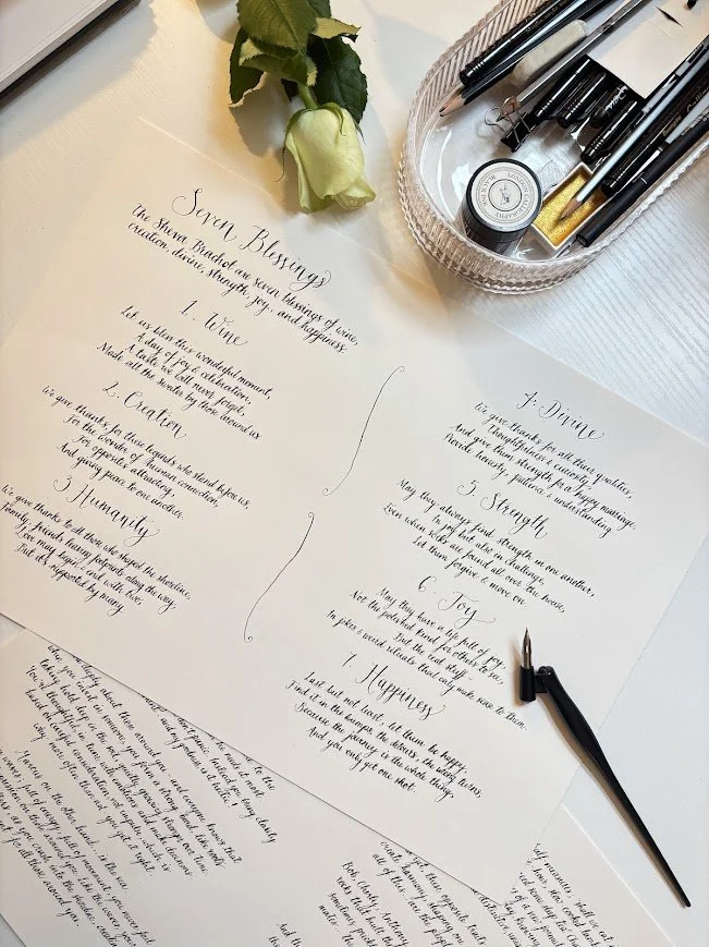 Handwritten calligraphy on white paper listing seven blessings with a pen, a rose, and a box of pens and ink on a white table.
