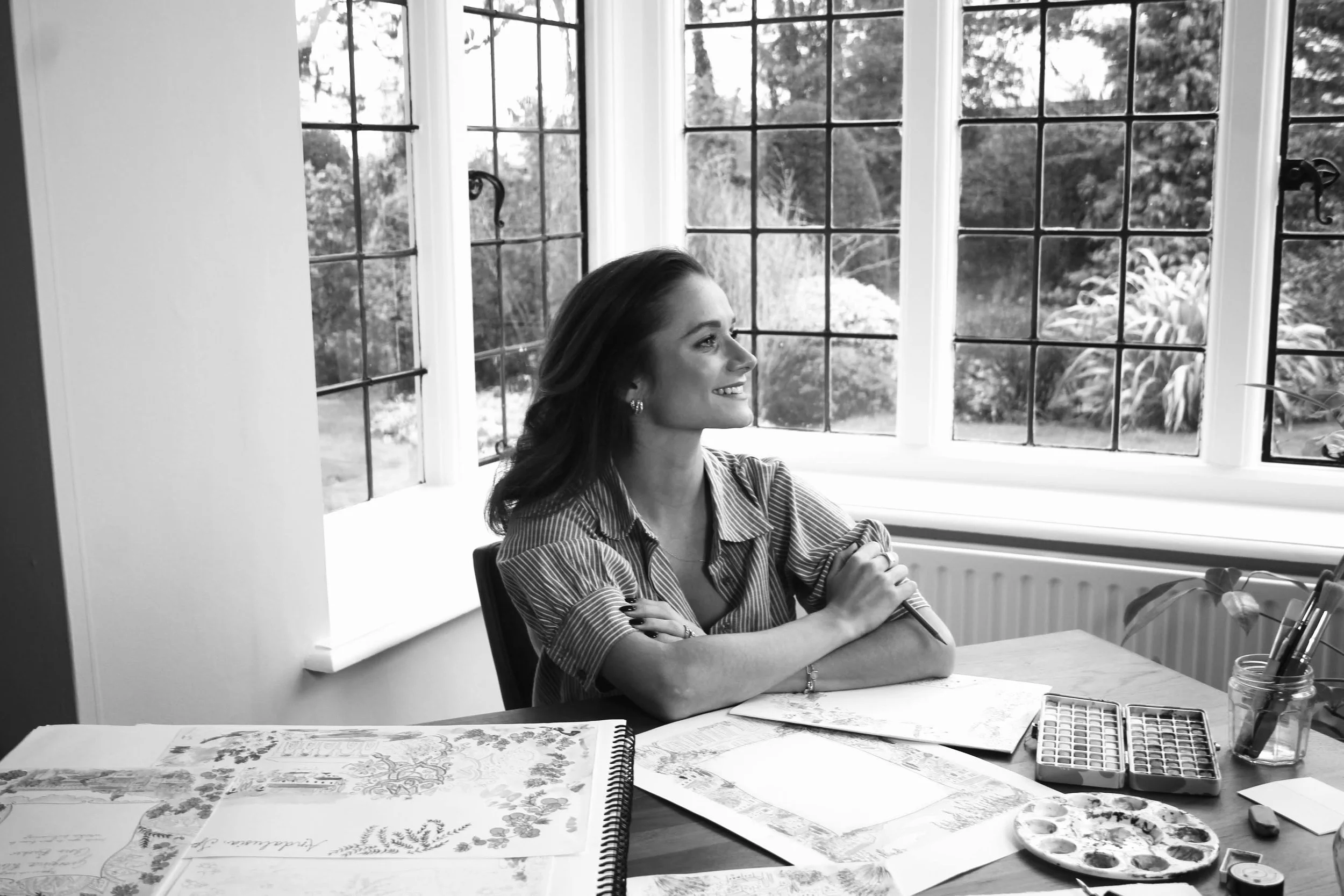 A woman sitting at a table by a large window, smiling and looking outside, surrounded by art supplies and sketches.