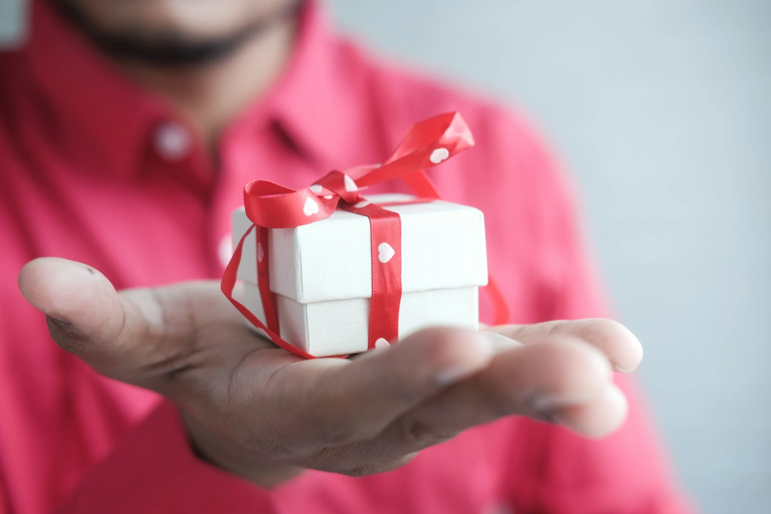 A person in a pink shirt holding a small white gift box with a red ribbon and bow