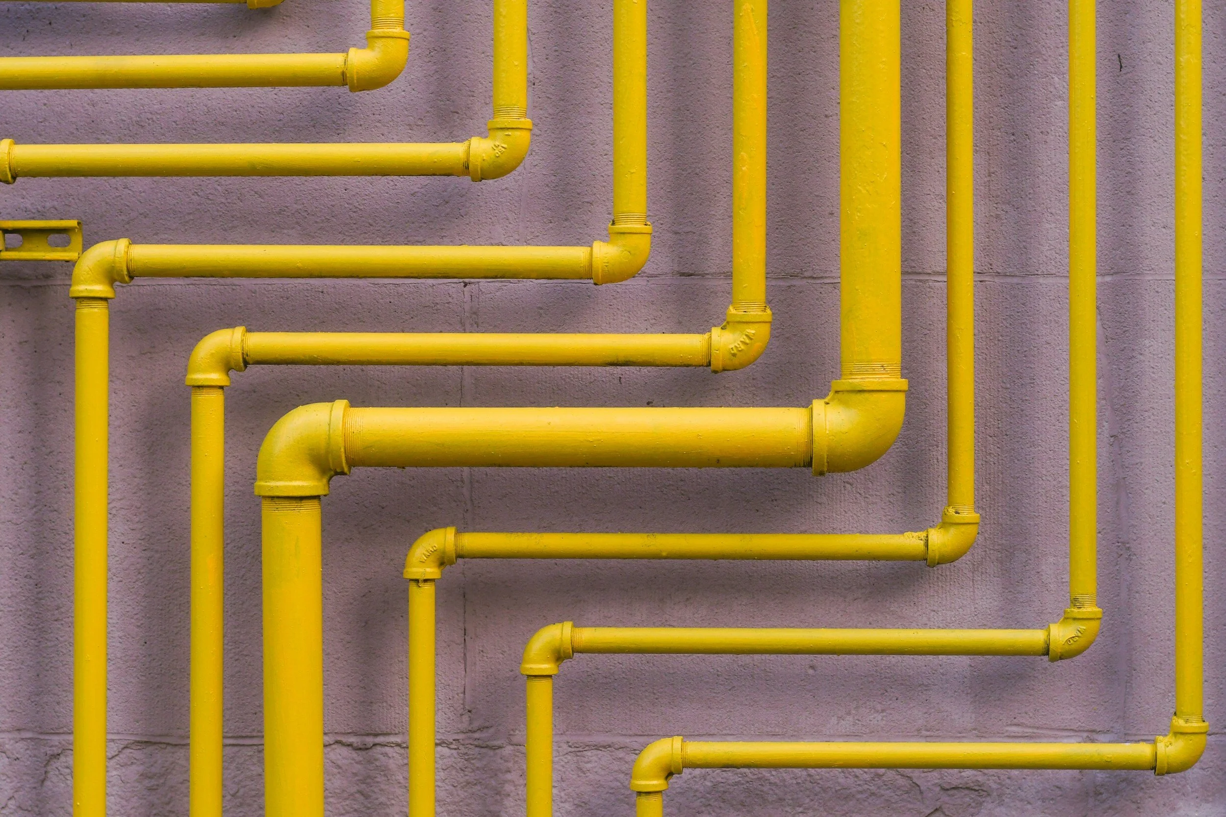 Multiple yellow pipes mounted on a gray wall, arranged in a pattern with some pipes bending and turning at right angles.