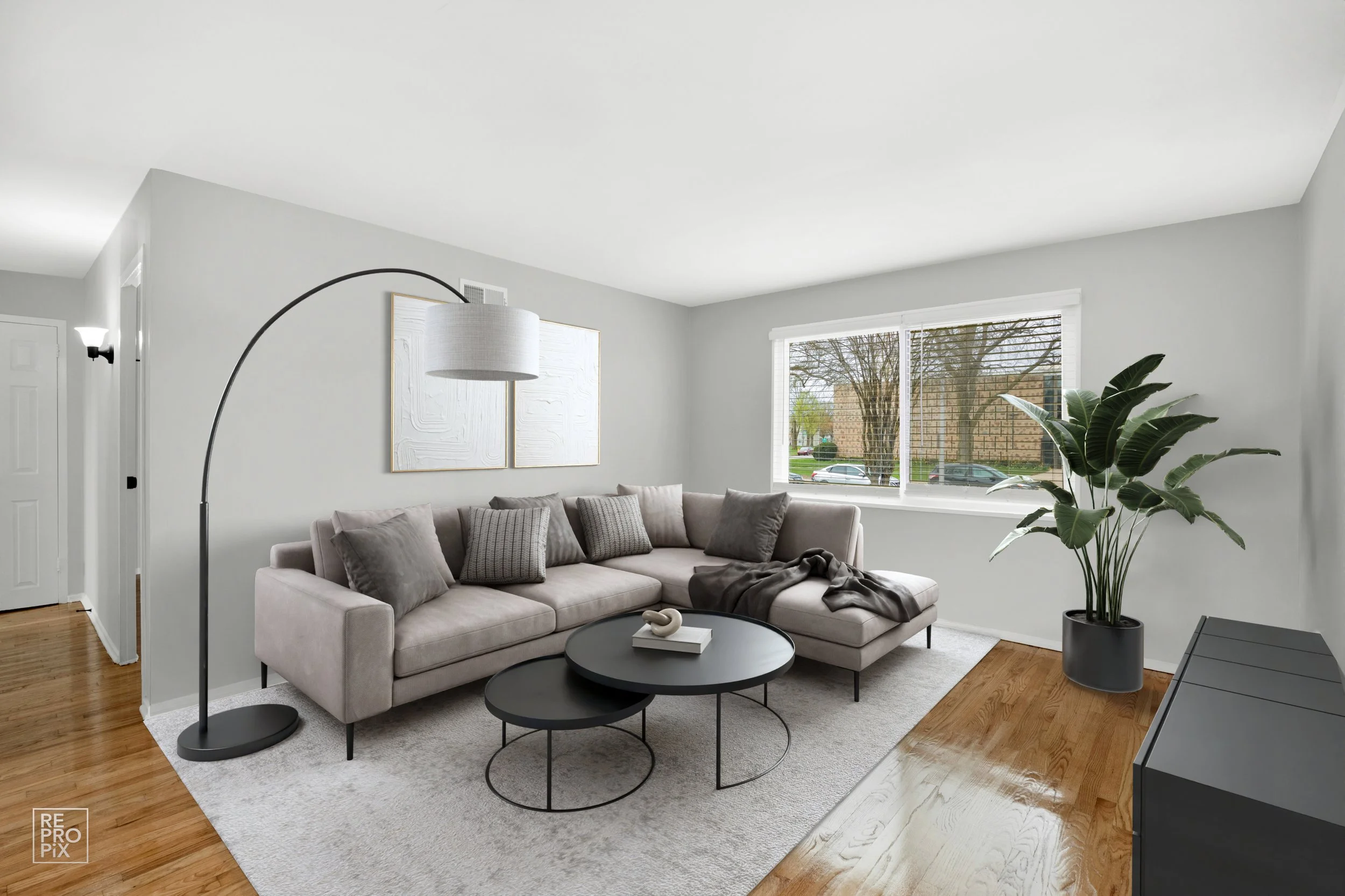Bright living room with a gray L-shaped sofa, gray and patterned pillows, a black arc floor lamp, and a large window with a tree view. Coffee tables and a potted plant are also visible.