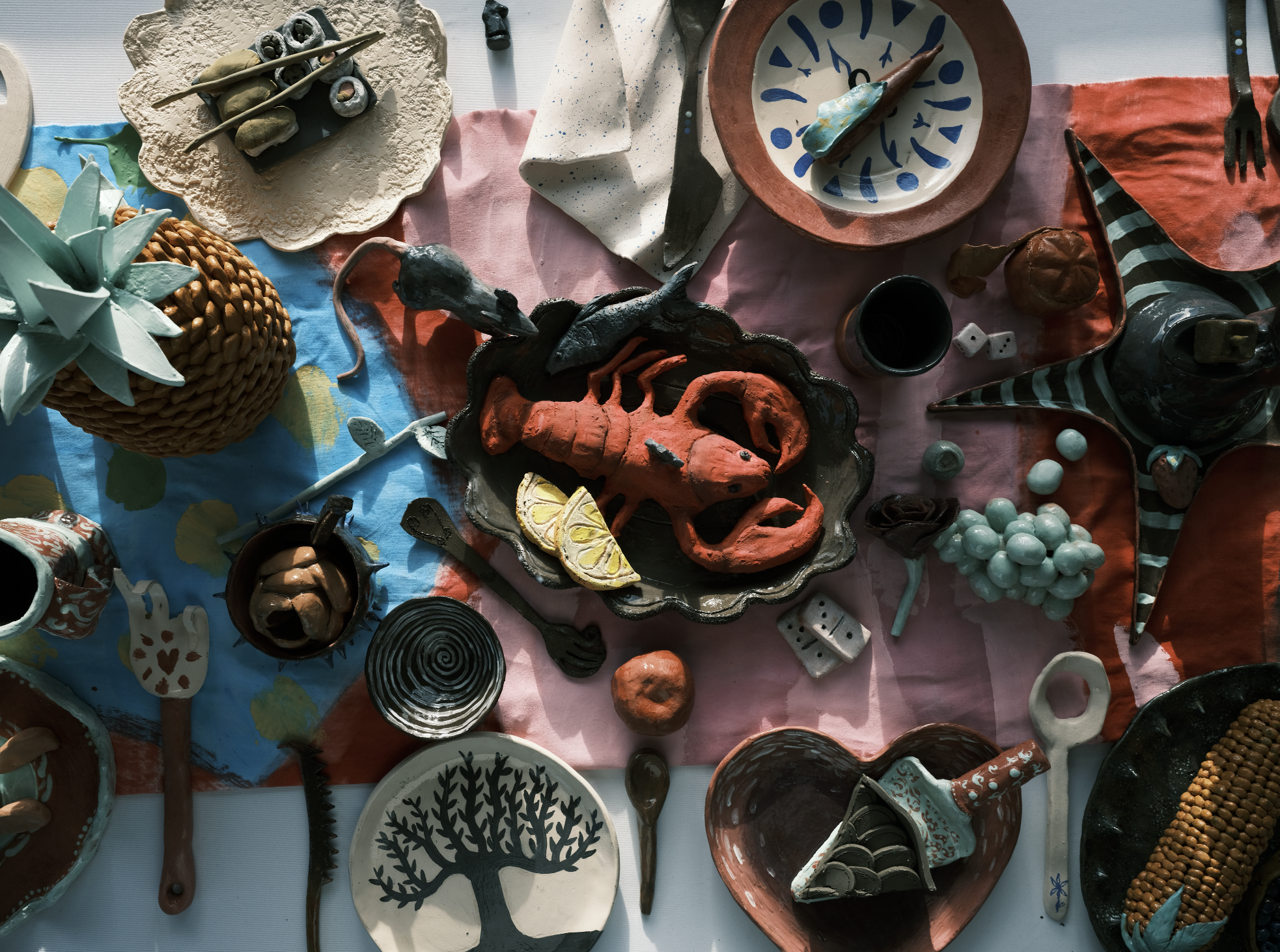 Table recouverte d'objets en céramique, comprenant un plat avec un homard, un ananas, une coupe noire avec un motif en spirale, une assiette avec un arbre noir, des figurines et autres objets décoratifs.
