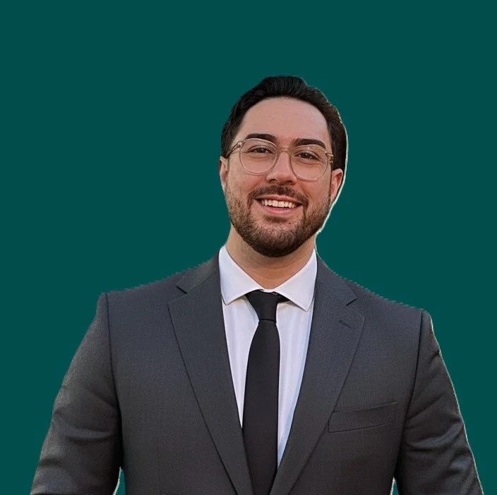 Portrait of a young man with light brown, curly hair, light skin, wearing a dark suit and tie, against a plain dark background.