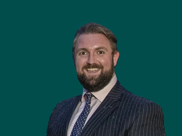 Michael Davie smiling, wearing a striped suit and a polka dot tie, against a dark background.