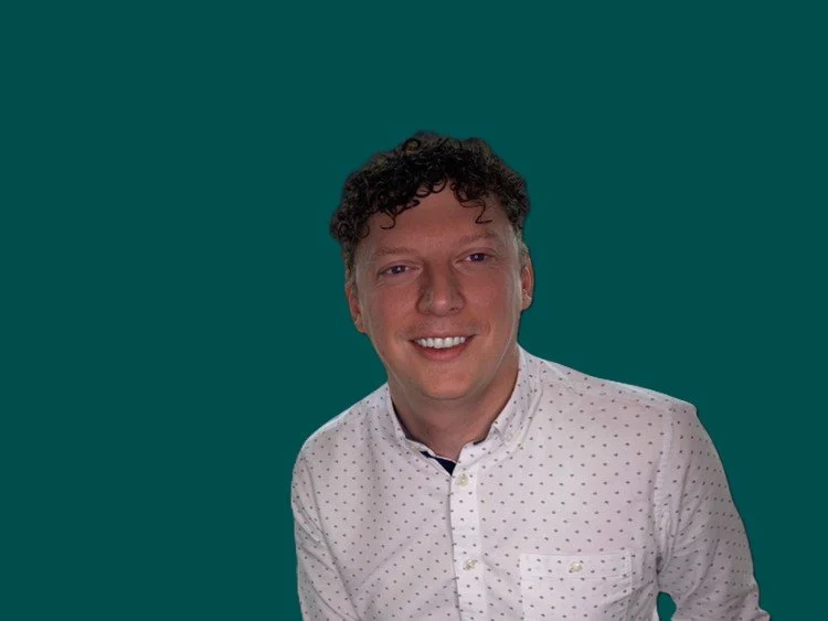 Headshot of a young man with curly dark hair smiling, wearing a white button-up shirt with small polka dots, against a solid dark background.