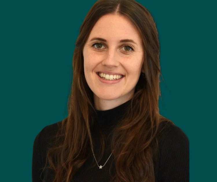 Portrait of a woman with long brown hair, wearing a black turtleneck and a star-shaped necklace, smiling against a dark background.