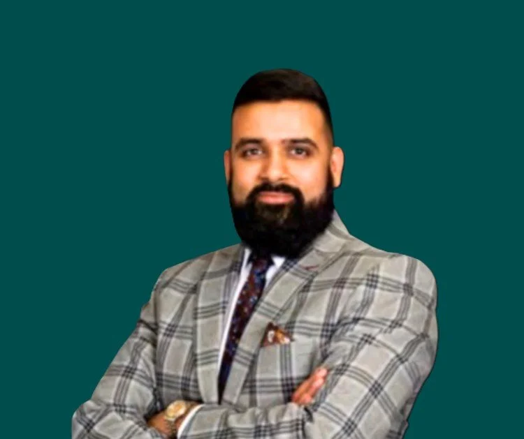 A man with a beard and short hair, wearing a checked blazer, white shirt, and tie, standing with crossed arms against a dark background.