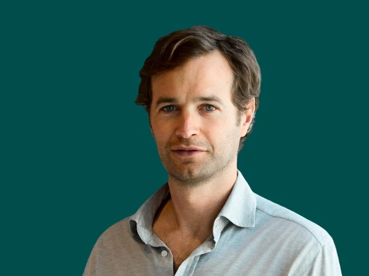 Liam Hurley wearing a light grey button-up shirt, is looking directly at the camera against a dark background.