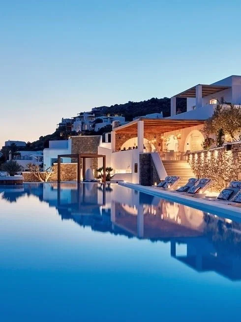 Luxury hotel with a large swimming pool reflecting the evening sky, white modern architecture, and hillside surroundings.