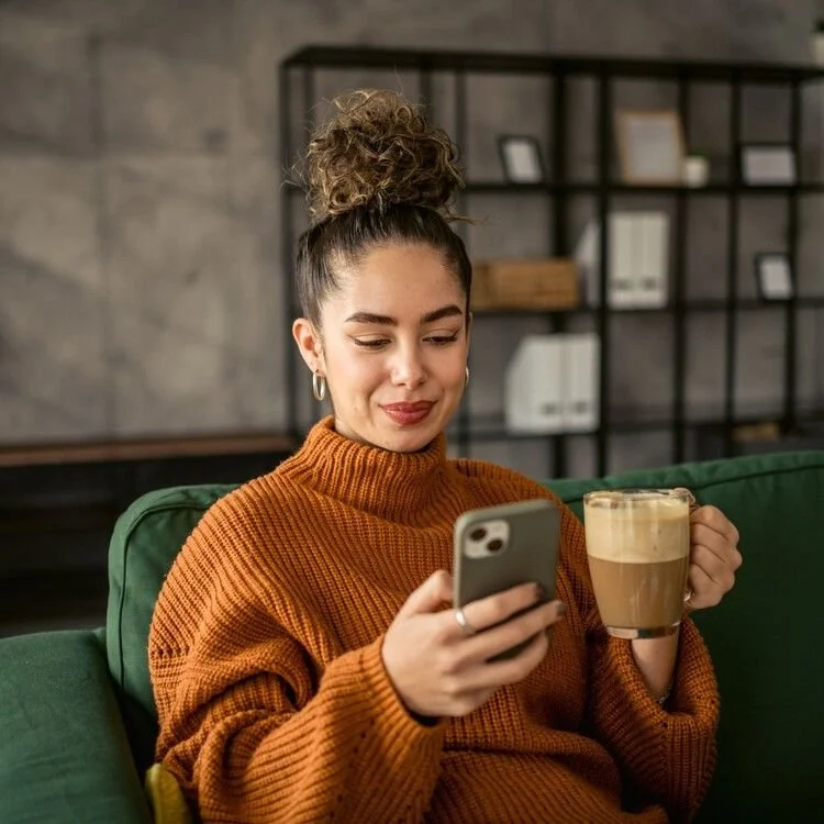 A young woman with curly hair in a messy bun, wearing a brown sweater, sitting on a green couch, holding a smartphone in her right hand and a mug of coffee in her left, smiling slightly in a modern living room setting.