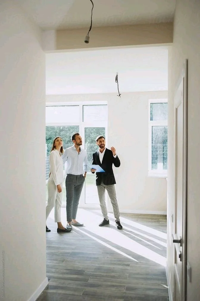 Three people looking at an empty room with large windows, sunlight streaming in, and a light fixture hanging from the ceiling, possibly during a property tour or inspection.