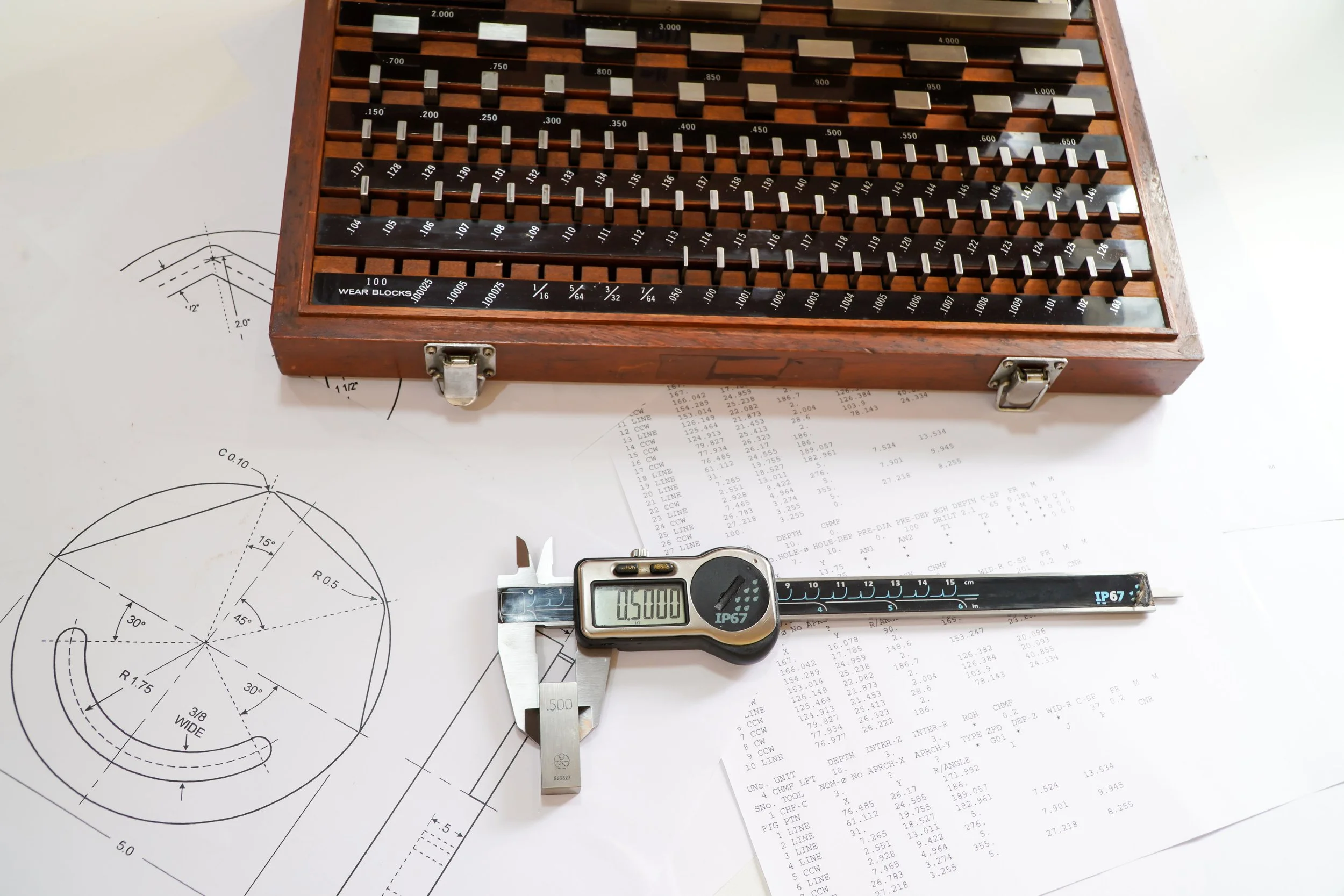 A detailed technical drawing of a mechanical part on white paper, with a digital caliper measuring the drawing, and a wooden box with metal clamps containing a set of small metal components on the surface.