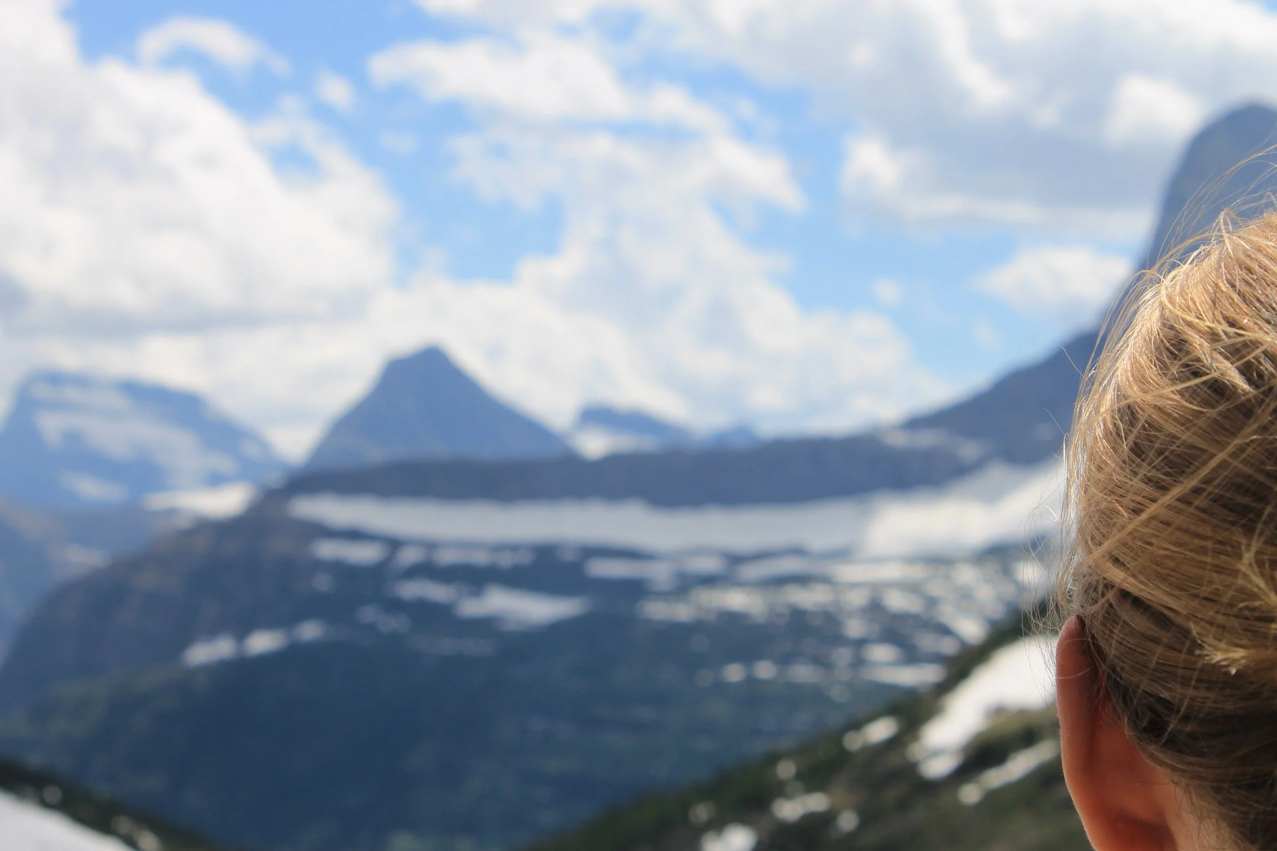 A person with blonde hair overlooking snow-capped mountains under a blue sky with clouds.