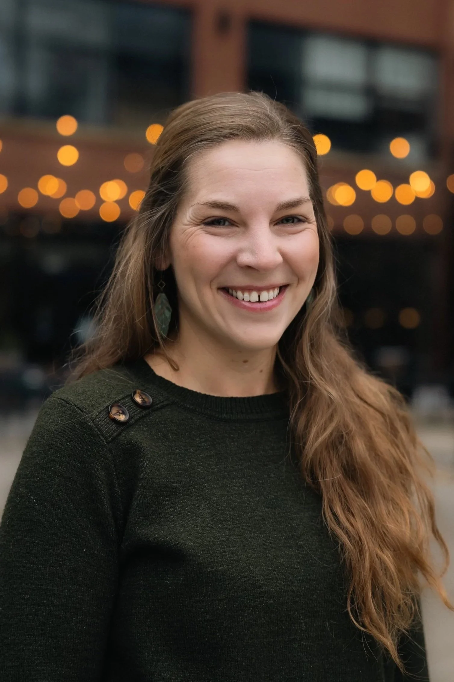 A woman with long, wavy brown hair smiling outdoors at night, wearing a dark sweater with button details on the shoulder, with bokeh lights in the background.