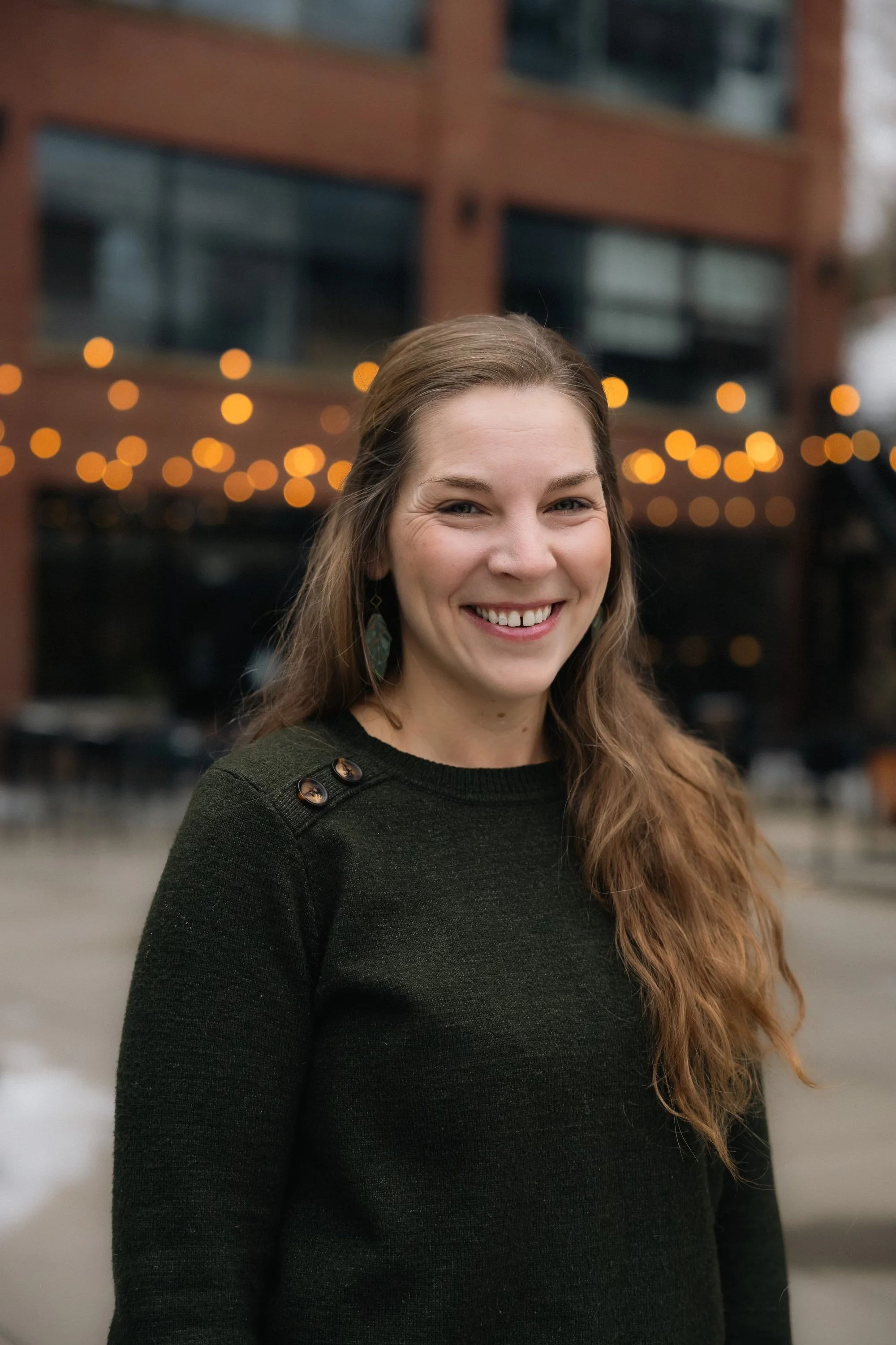 A smiling woman with long, wavy brown hair and teal earrings standing outdoors in front of a building with lit string lights.
