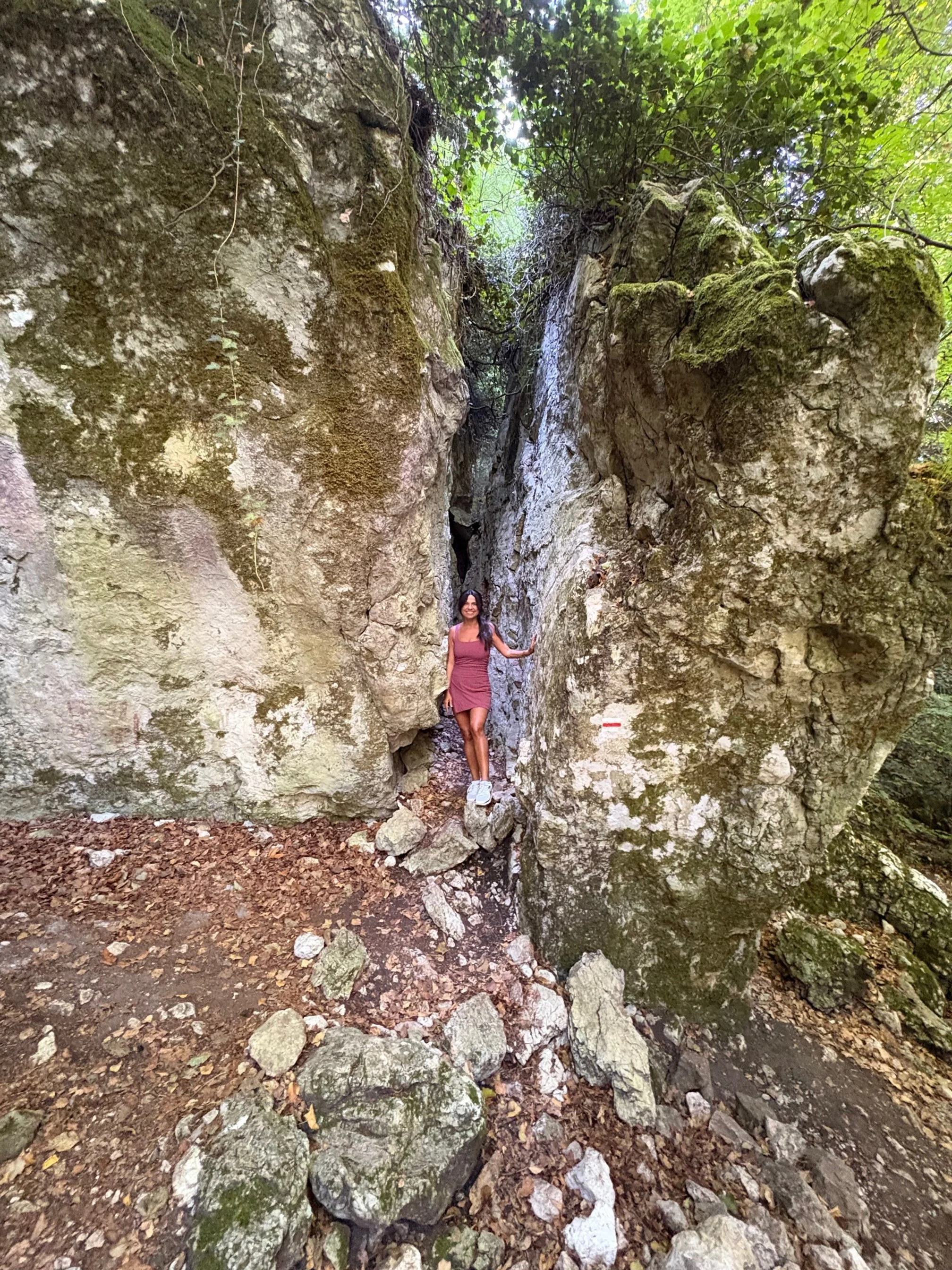 Une femme en robe rose clair et baskets blanches se tient entre deux grandes roches dans une forêt verte, en marchant sur un chemin de terre et de rochers.