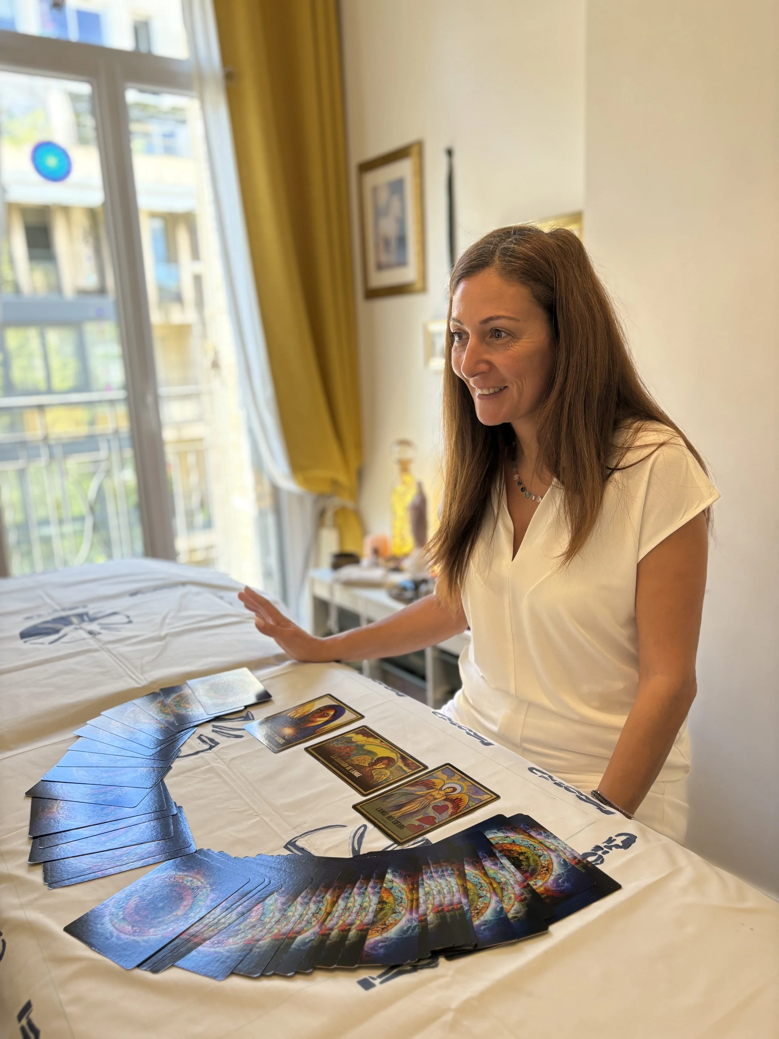 Une femme souriante assise à une table, faisant la lecture de cartes de tarot disposées en arc sur la table, dans une pièce lumineuse avec paysages visibles à travers une fenêtre.
