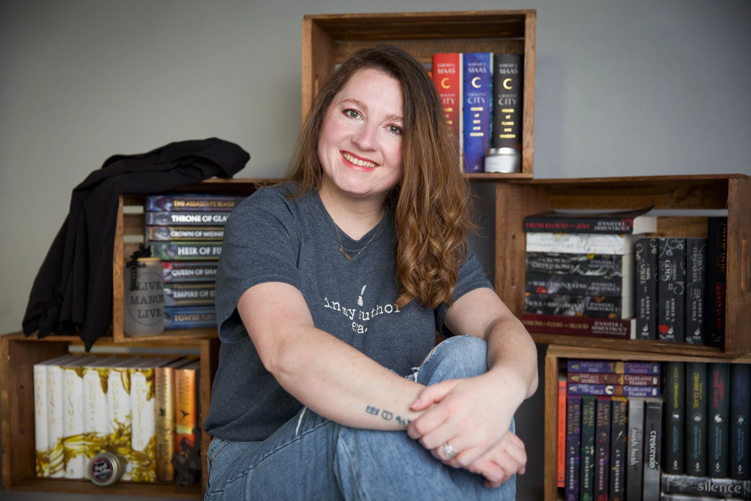 A young woman with long brown hair and fair skin sitting in front of a bookshelf filled with colorful books. She is smiling, wearing a dark gray T-shirt and ripped jeans, with tattoos visible on her left arm.