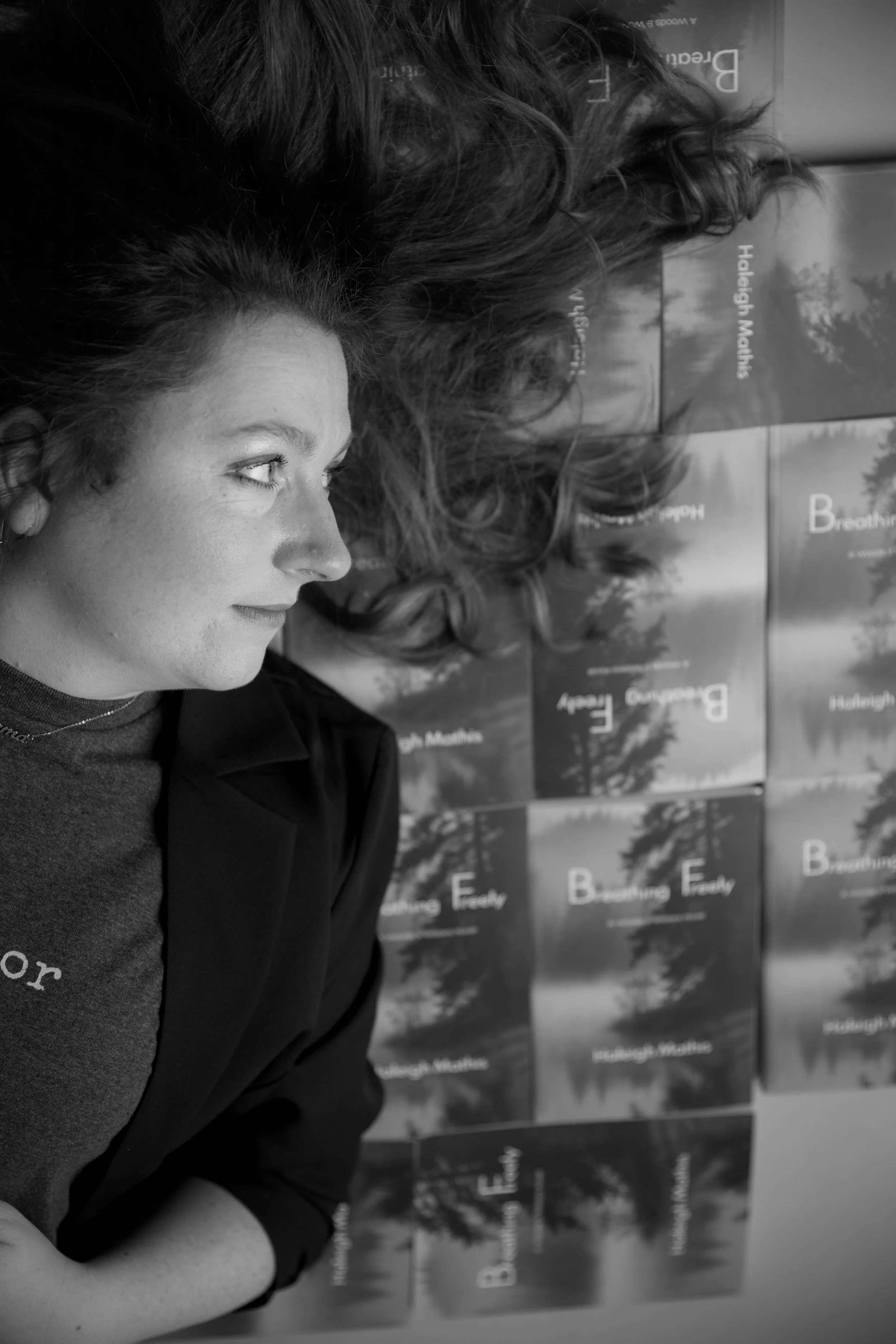 Side profile of a woman with curly hair looking at a wall of magazines or books with titles like 'Breathing Easy' and 'Helaleigh Malkins'. The image is in black and white.
