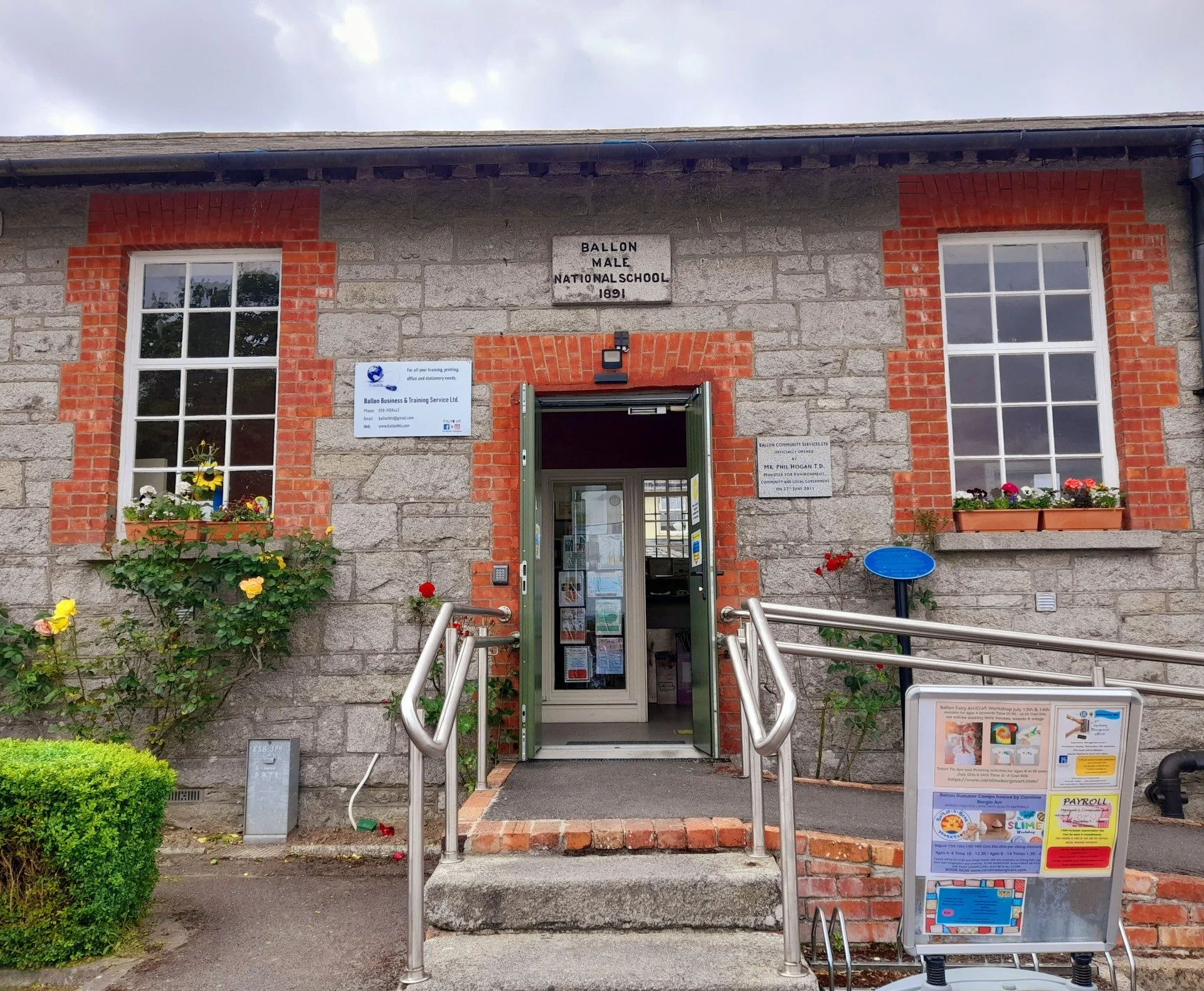 Front view of a stone and brick school building with two large windows, flower boxes, a sign reading 'Ballon Male National School 1891', and an open door with a ramp and stairs leading up to it.