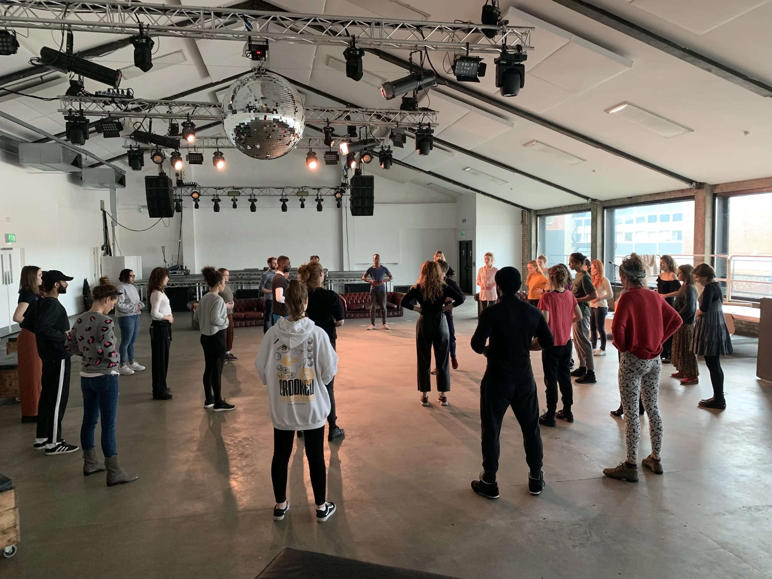 A group of people at a workshop stands in a circle in a spacious, well-lit room with large windows and a ceiling with lighting and sound equipment. There is a disco ball hanging from the ceiling.